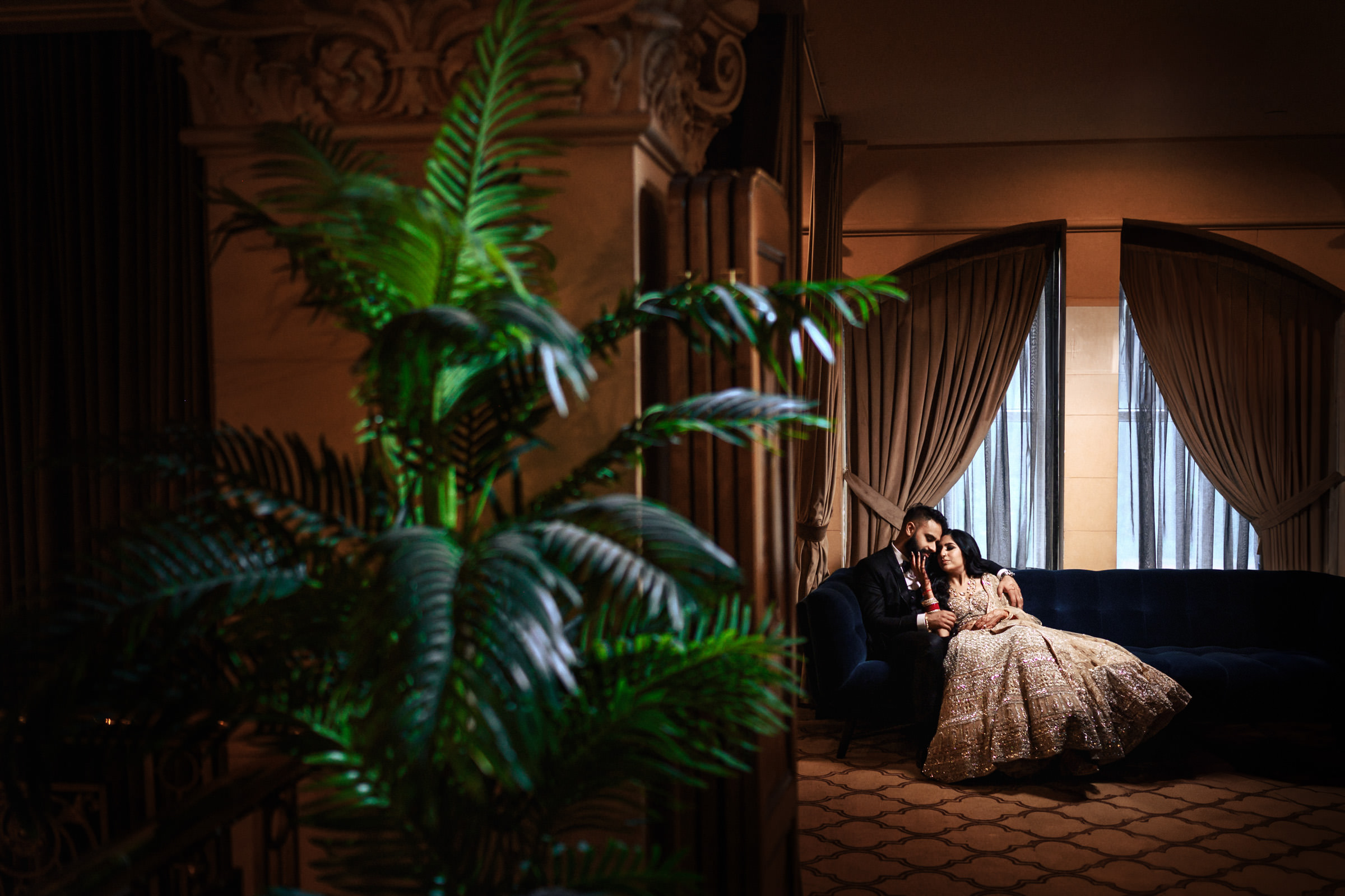 A couple dressed in traditional attire sits closely together on a dark blue couch in an elegantly decorated room with beige walls and tall windows with curtains. A green fern partially obscures the view on the left, adding a touch of nature to the scene.