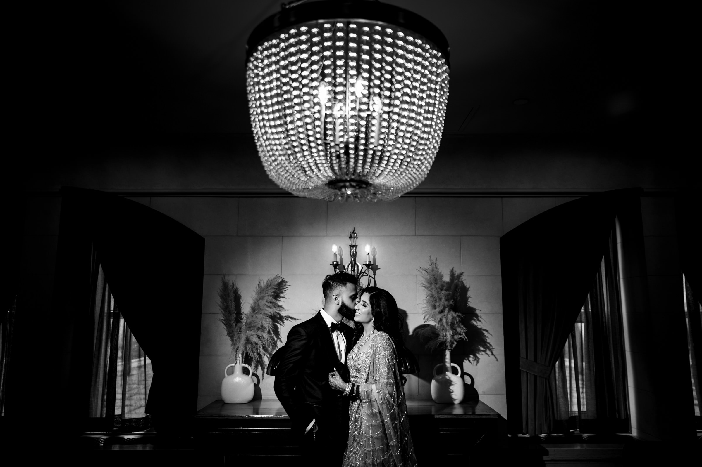 A black and white photograph of a couple standing close together in an elegant room. Above them is a large, ornate chandelier. Both are dressed formally; the man in a tuxedo and the woman in a detailed dress. The background features vases and curtains.