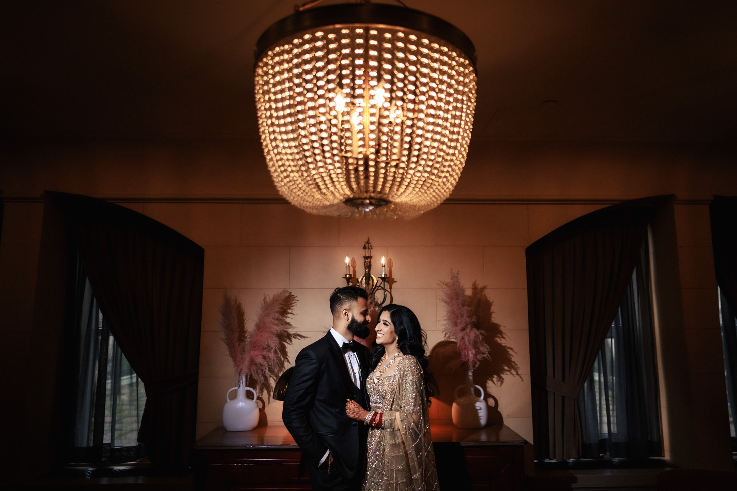 A couple stands under a large, ornate chandelier in a dimly lit room. They are dressed in formal attire, with the woman in an embellished gown and the man in a black suit. Behind them are decorative pieces and tall, dark drapes covering windows.