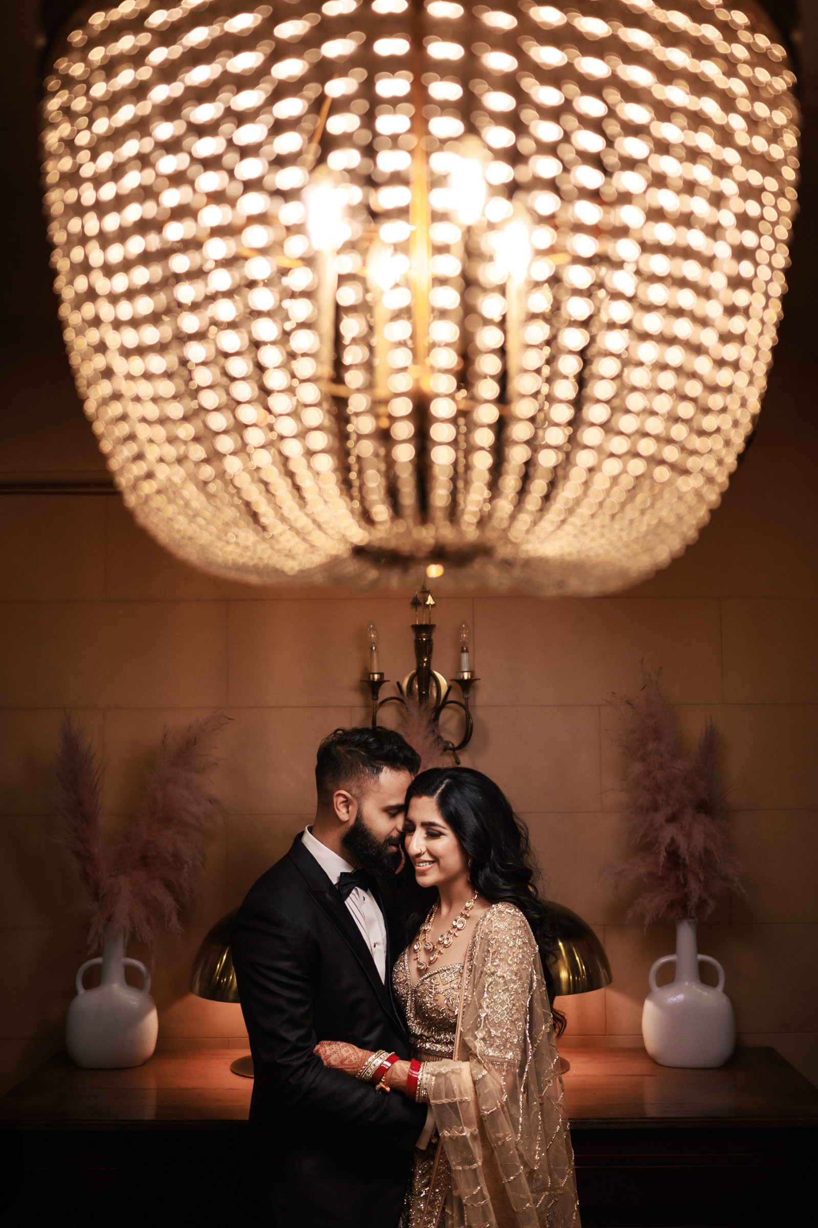 A couple dressed elegantly stands close under a large, ornate chandelier. The man wears a black tuxedo, and the woman is in a golden, embellished traditional outfit. They are indoors, smiling at each other with decorative flowers and a lamp in the background.