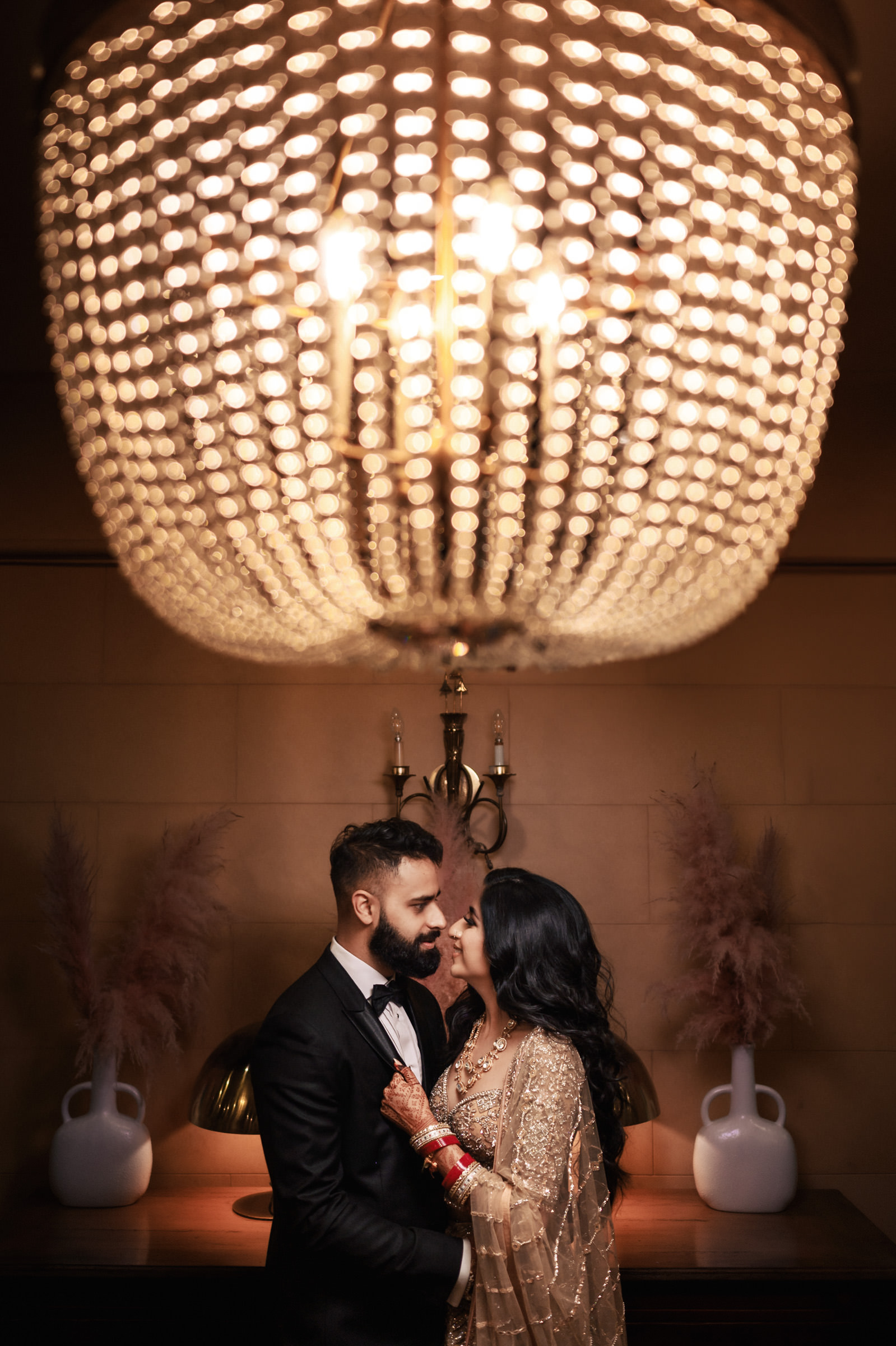 A couple gazing at each other under a sparkling chandelier. The man is in a black suit, and the woman in a traditional, embellished gown with red bridal bangles. The background features a wooden console with decorative vases and soft lighting.