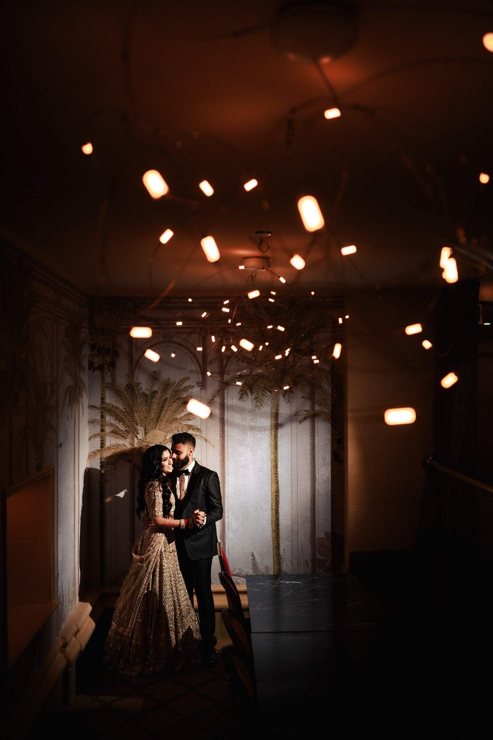 A couple dressed in formal attire embraces under warm, ambient lighting. The woman wears a detailed, flowing gown, while the man dons a suit. They stand in a dimly lit room with decorative wallpaper featuring tree motifs and hanging lights, creating a romantic atmosphere.