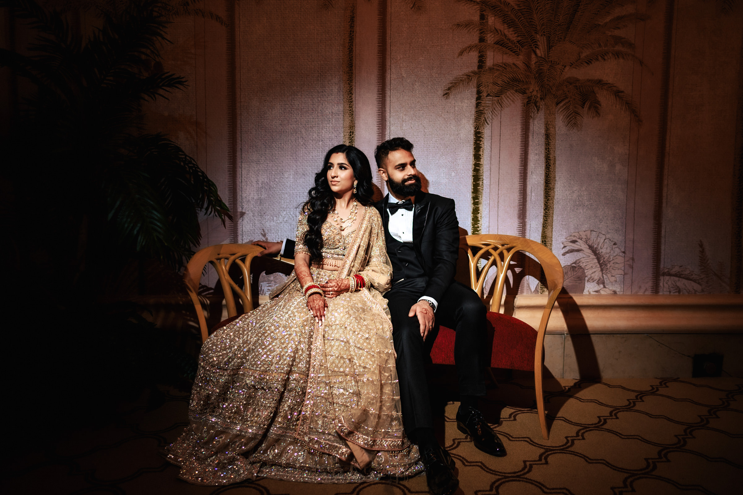 A couple sits on a red bench in front of a backdrop featuring palm trees. The woman is dressed in an intricate beige lehenga and the man is wearing a black suit with a bow tie. Both appear to be posing for a formal photograph.