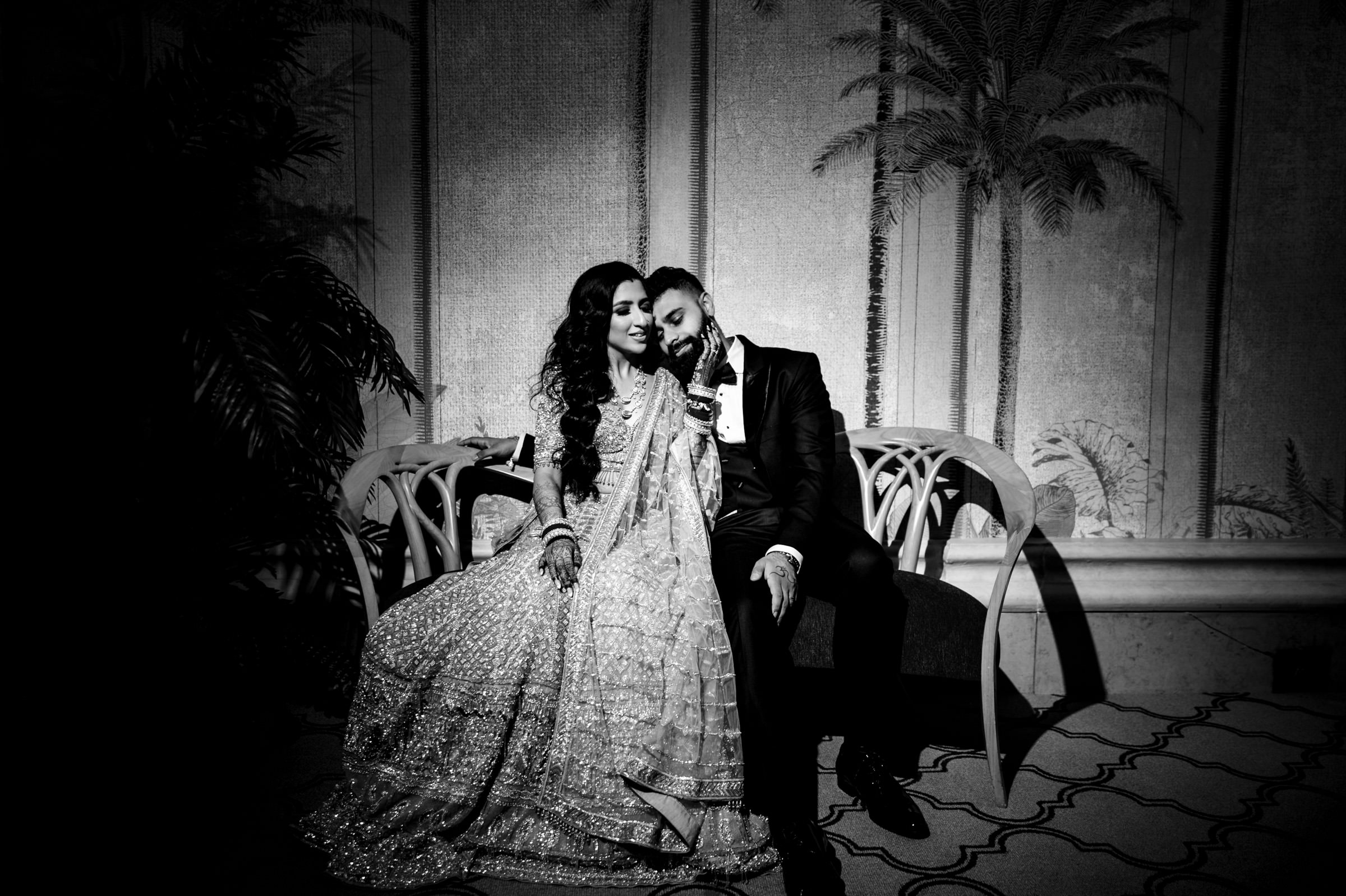 A black and white photo of a couple sitting closely together on an ornate bench. The woman is wearing an embellished traditional dress and rests her head on the man's shoulder. The man is wearing a suit and is gently kissing her head. Palm trees are painted on the backdrop.