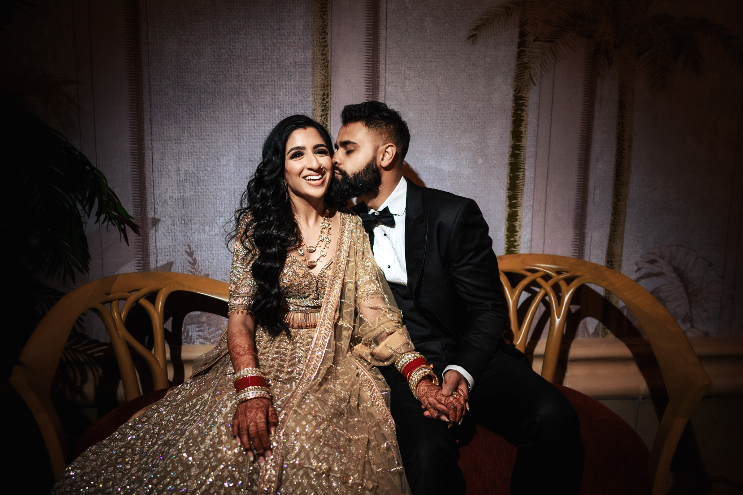 A couple is seated on a wooden bench, holding hands. The woman, dressed in an ornate traditional outfit with intricate designs and red bangles, smiles. The man, in a black tuxedo, kisses her on the cheek. The background features subtle, decorative elements.