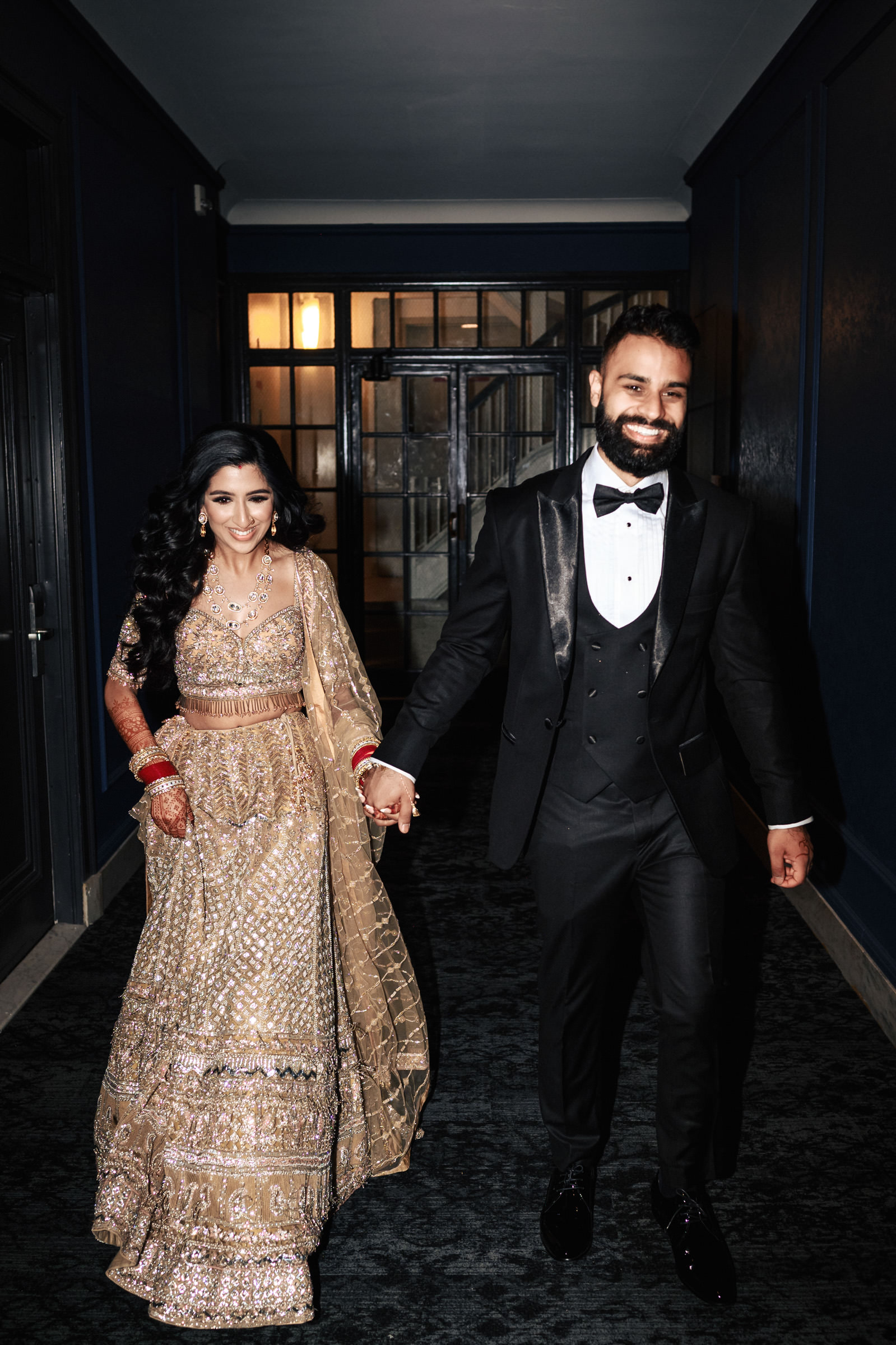 A couple is seen walking hand in hand down a hallway. The woman is wearing an ornate, traditional South Asian outfit, complete with jewelry and intricate embroidery, while the man is dressed in a black tuxedo. Both are smiling under the dim hallway lighting.