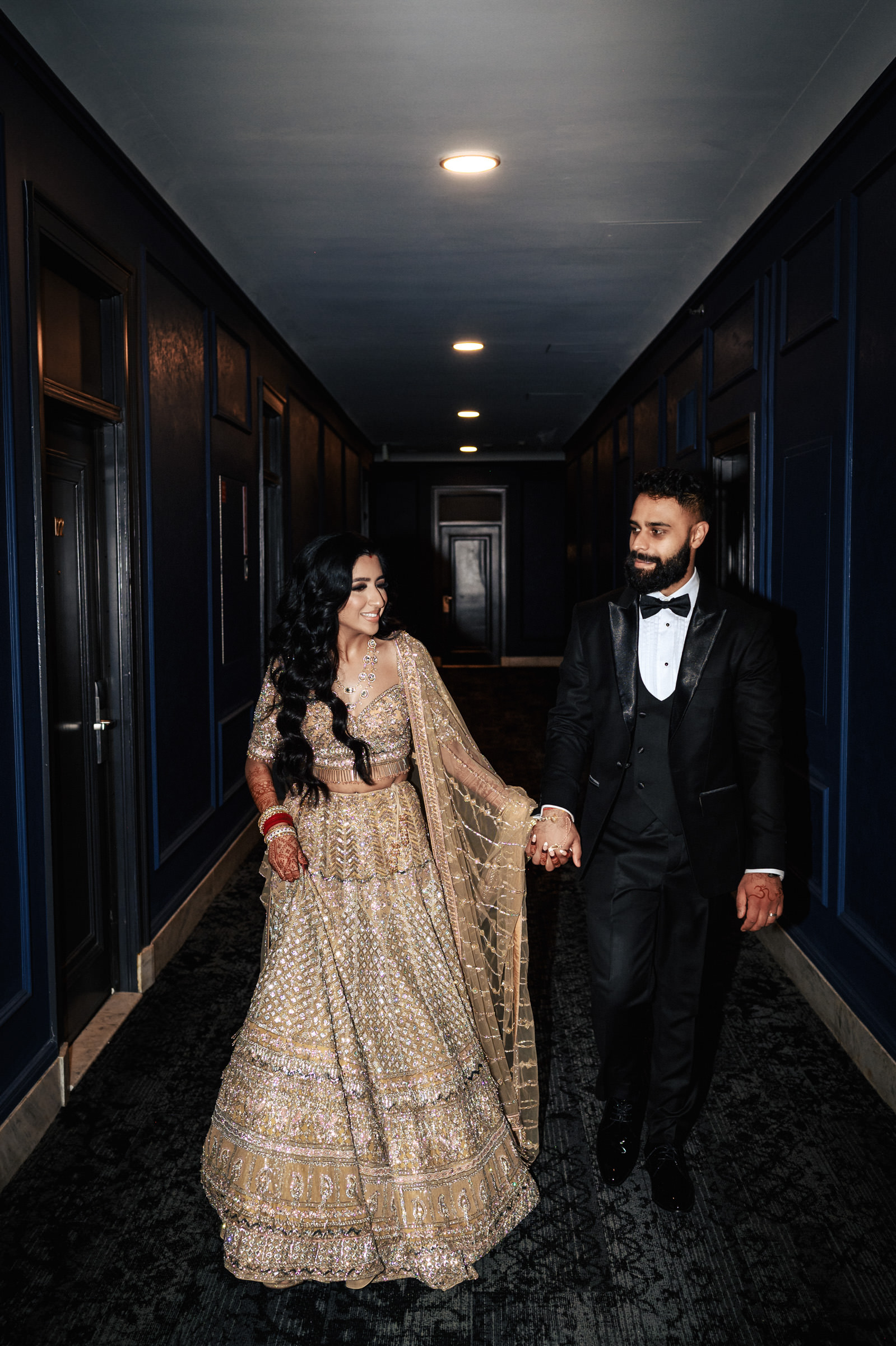 A couple, dressed in traditional Indian attire, walks hand-in-hand in a hallway. The woman wears an intricate beige lehenga adorned with jewelry, while the man sports a black tuxedo. Both are smiling warmly at each other, illuminated by overhead lights.
