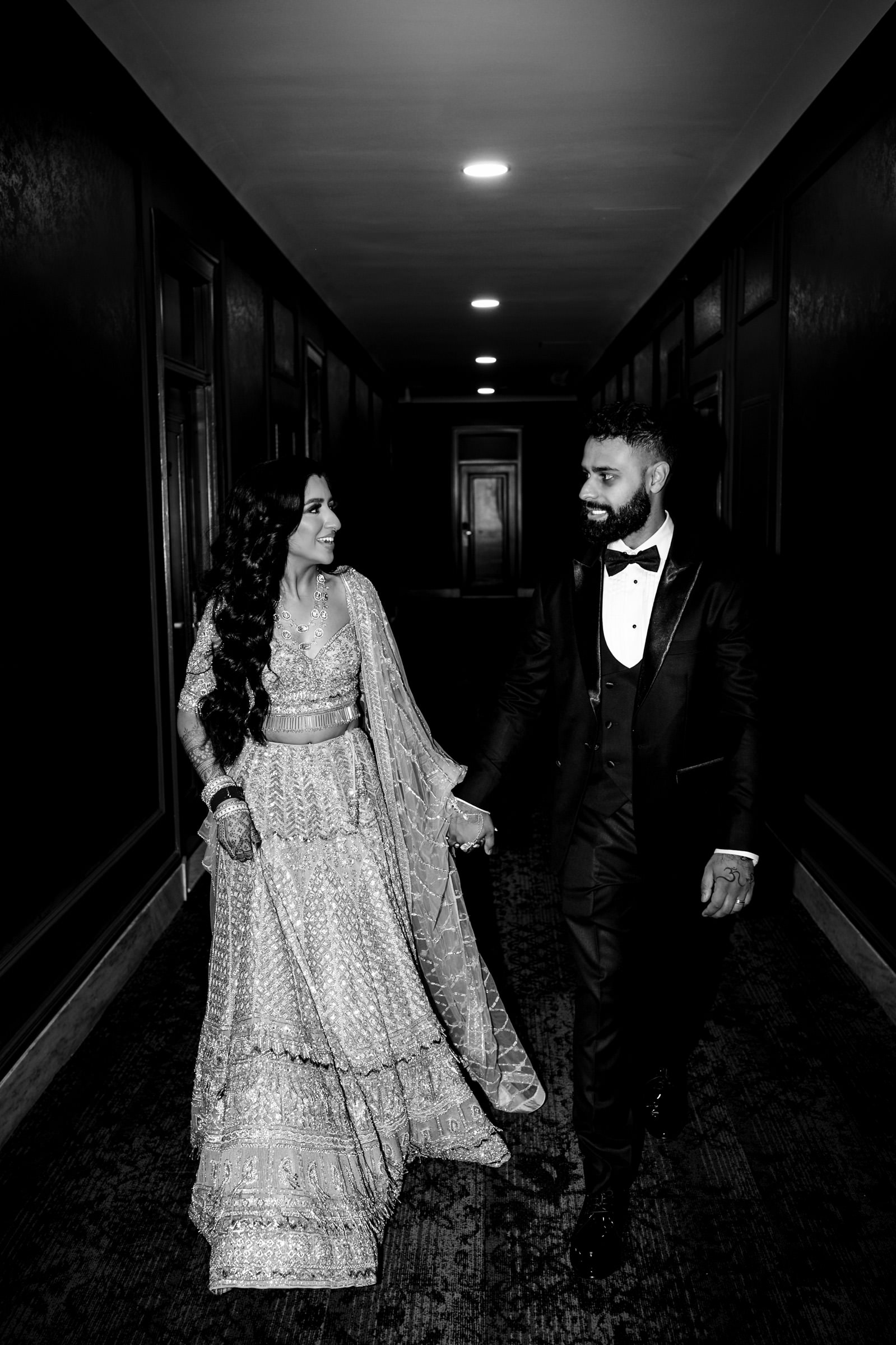 A couple in formal attire walk hand-in-hand down a corridor lined with framed panels. The woman wears a traditional, embellished outfit, and the man is in a classic black tuxedo. Both are smiling and looking at each other, illuminated by overhead lights.