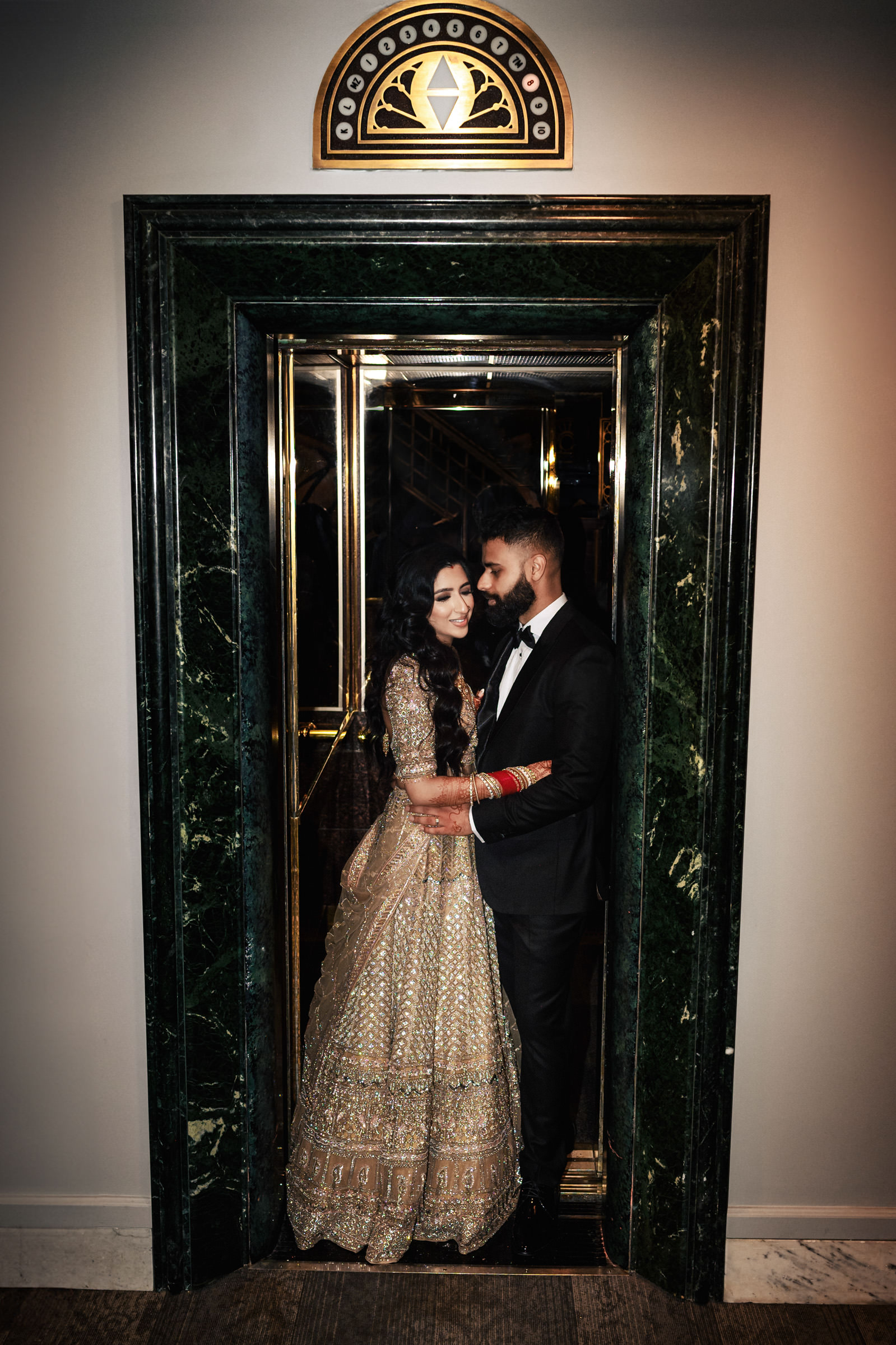 A couple is standing closely together in an ornate, marble-framed elevator. The woman is wearing an intricately designed traditional dress, and the man is dressed in a black suit. They both appear to be sharing an intimate moment, gazing at each other affectionately.