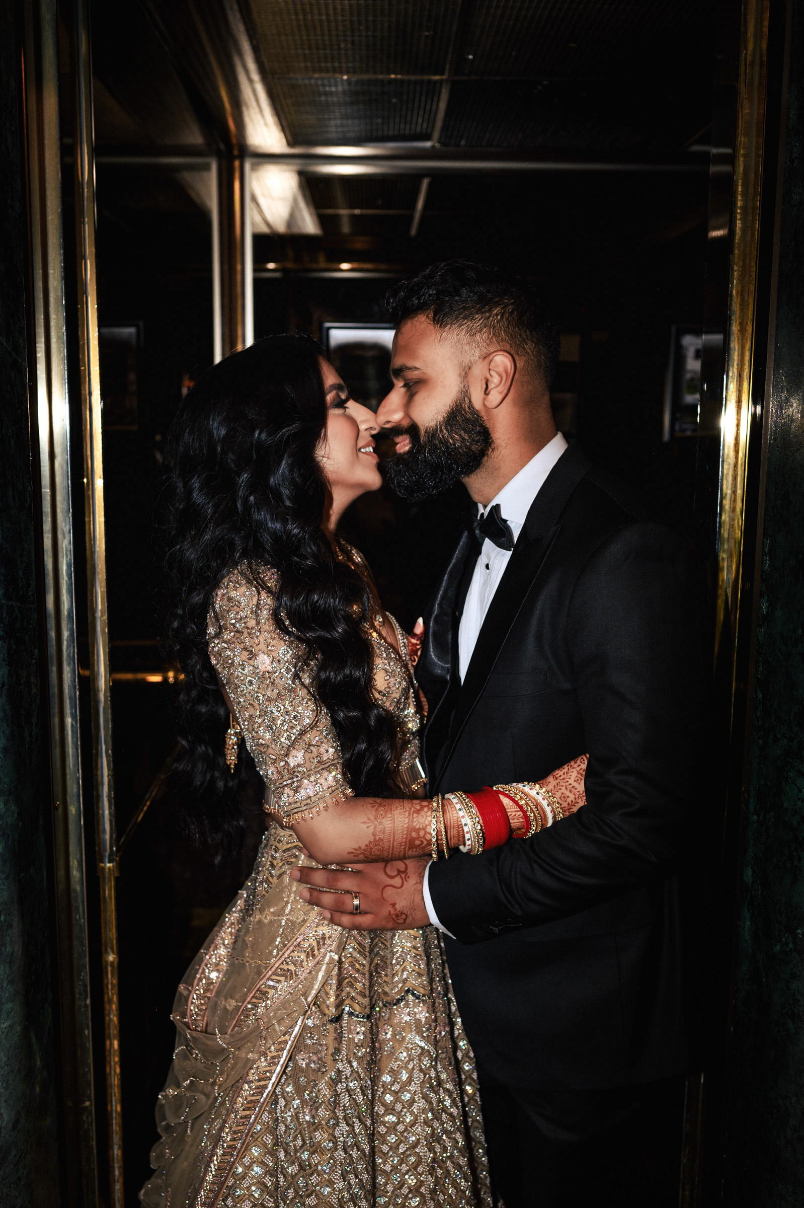 A couple stands in a close embrace, gazing into each other's eyes while dressed in formal attire. The woman wears an ornate, gold-colored gown with intricate details and red bangles, and the man is in a black tuxedo with a black bow tie.