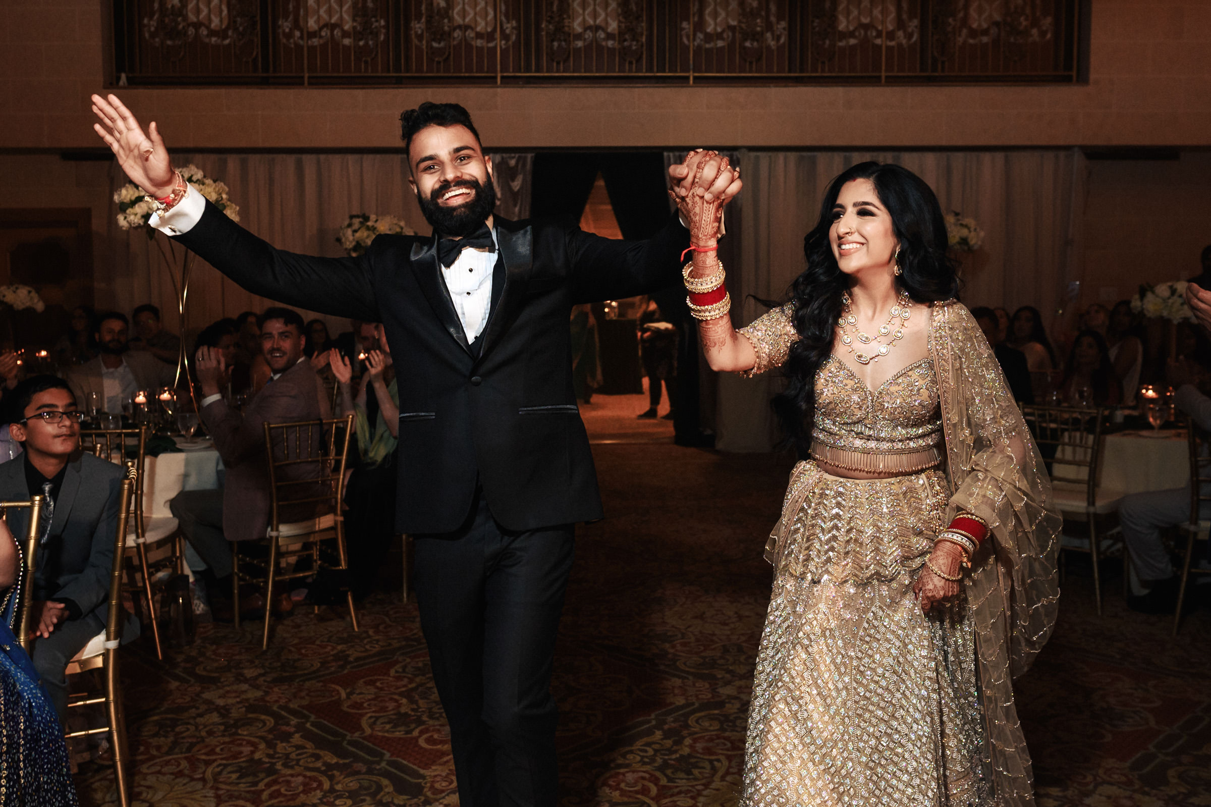 A couple in formal attire celebrates at a wedding reception. The groom wears a black suit and bow tie, while the bride dons a shimmering gold lehenga. They smile and hold hands, walking through a room filled with guests seated at decorated tables.