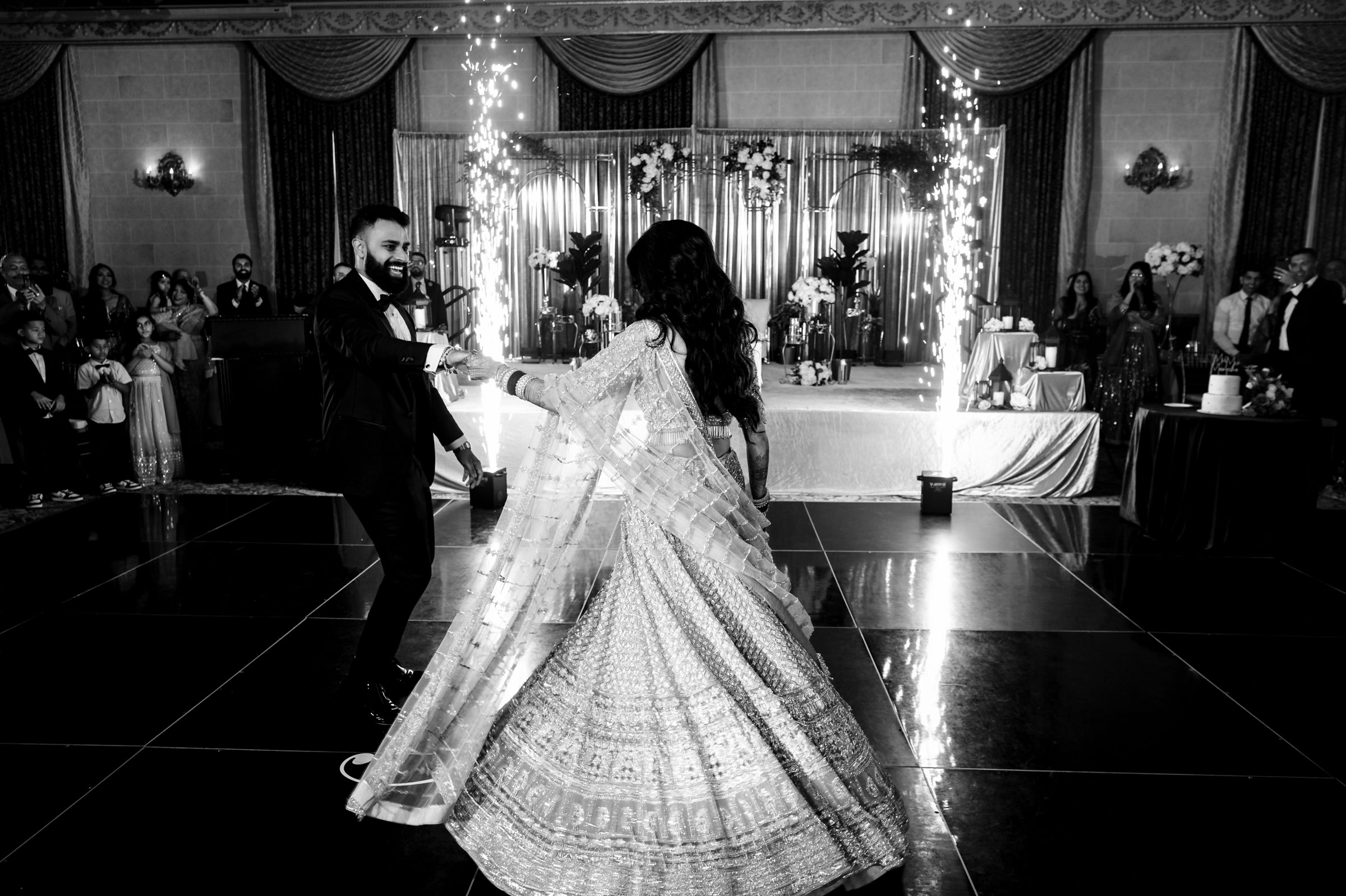 A couple dances in an elegant ballroom, with the woman in a detailed, traditional dress and the man in a suit. Guests stand in the background, and the room is adorned with drapes, flowers, and sparkling decorations.