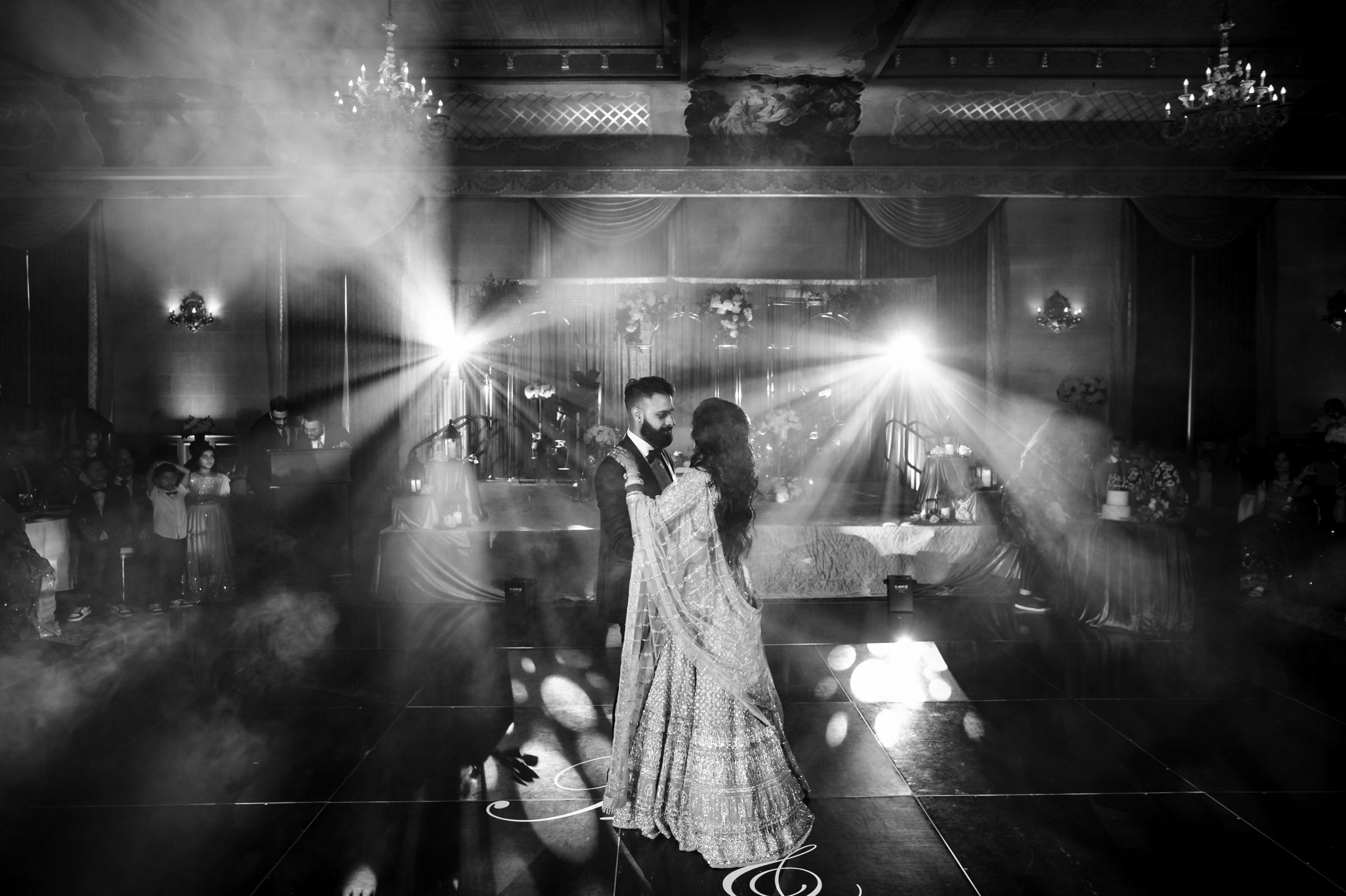 A couple dances in the center of a dimly lit ballroom adorned with chandeliers. Spotlights cast beams of light around them, creating a dramatic effect with mist enveloping the scene. The couple appears to be sharing an intimate moment on a glossy dance floor.