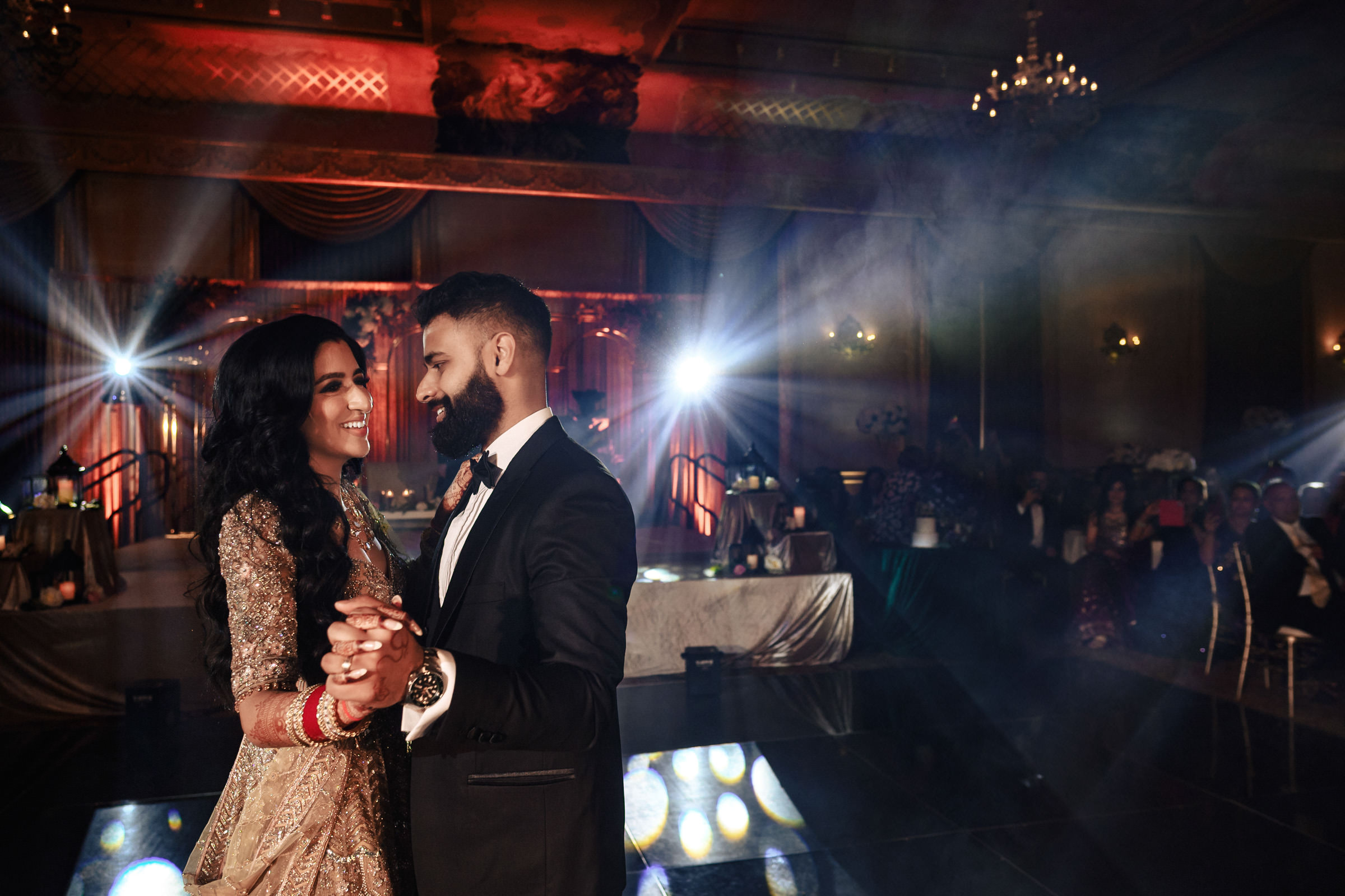 A couple, dressed in formal attire, dances together in a dimly lit, elegantly decorated hall. The woman wears a gold and red dress, the man a black suit. Bright lights illuminate the background, highlighting their joyful expressions. Guests sit at tables, observing the dance.