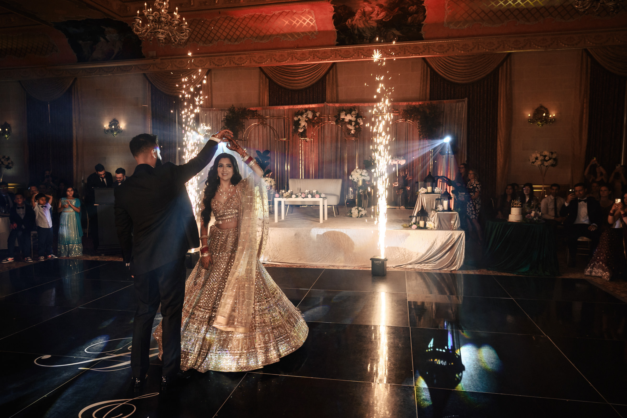 A bride and groom dance in an opulent hall, illuminated by sparklers. The bride wears a shimmering lehenga while the groom is in a black suit. The background features decorative drapes, flowers, and a cake on a table, with guests watching them.