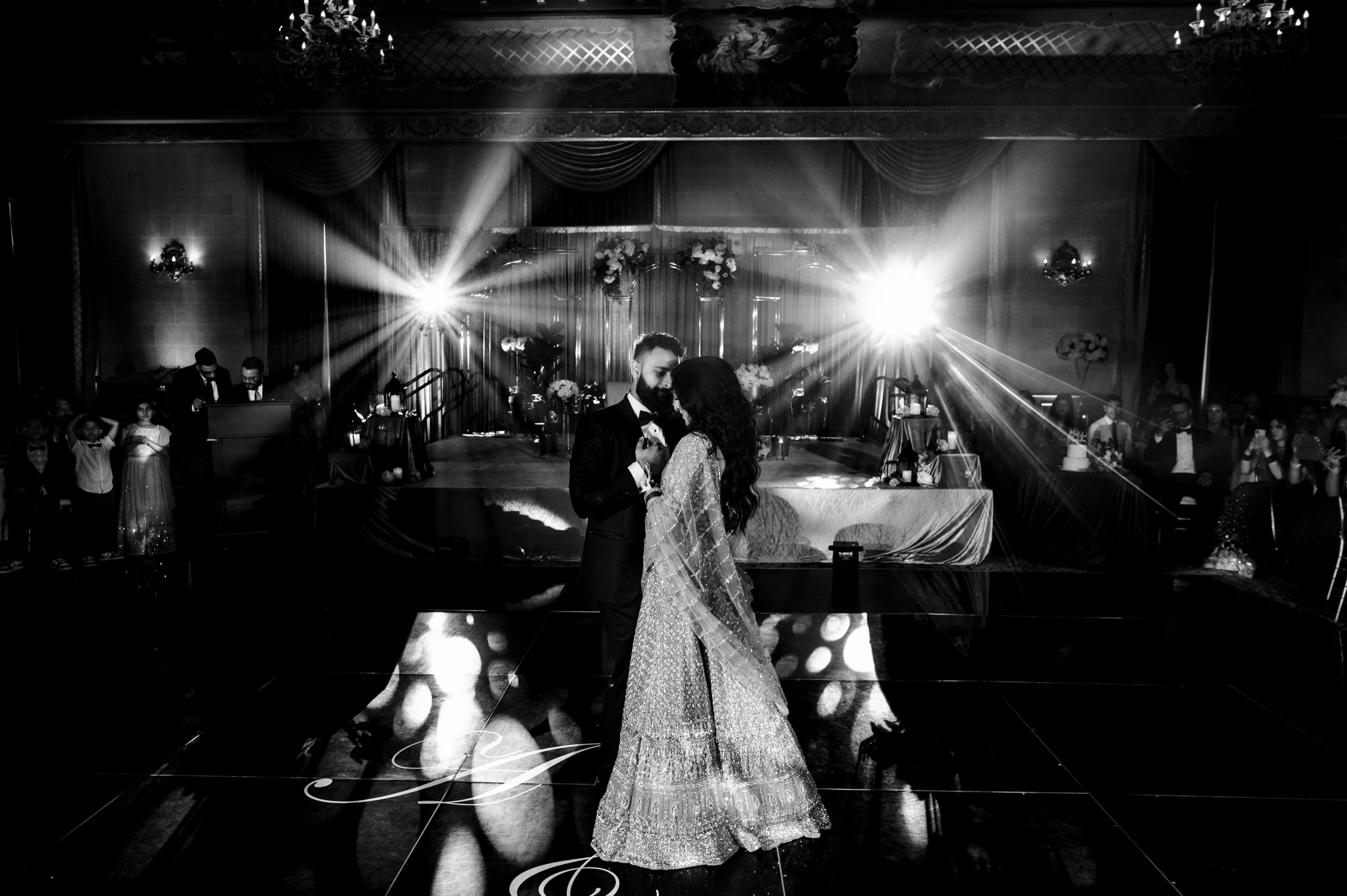 A couple dances together in an elegant ballroom adorned with chandeliers and drapery. They are illuminated by bright stage lights, creating a romantic atmosphere. The bride wears a detailed gown, and the groom is in a suit. Guests watch from the background.