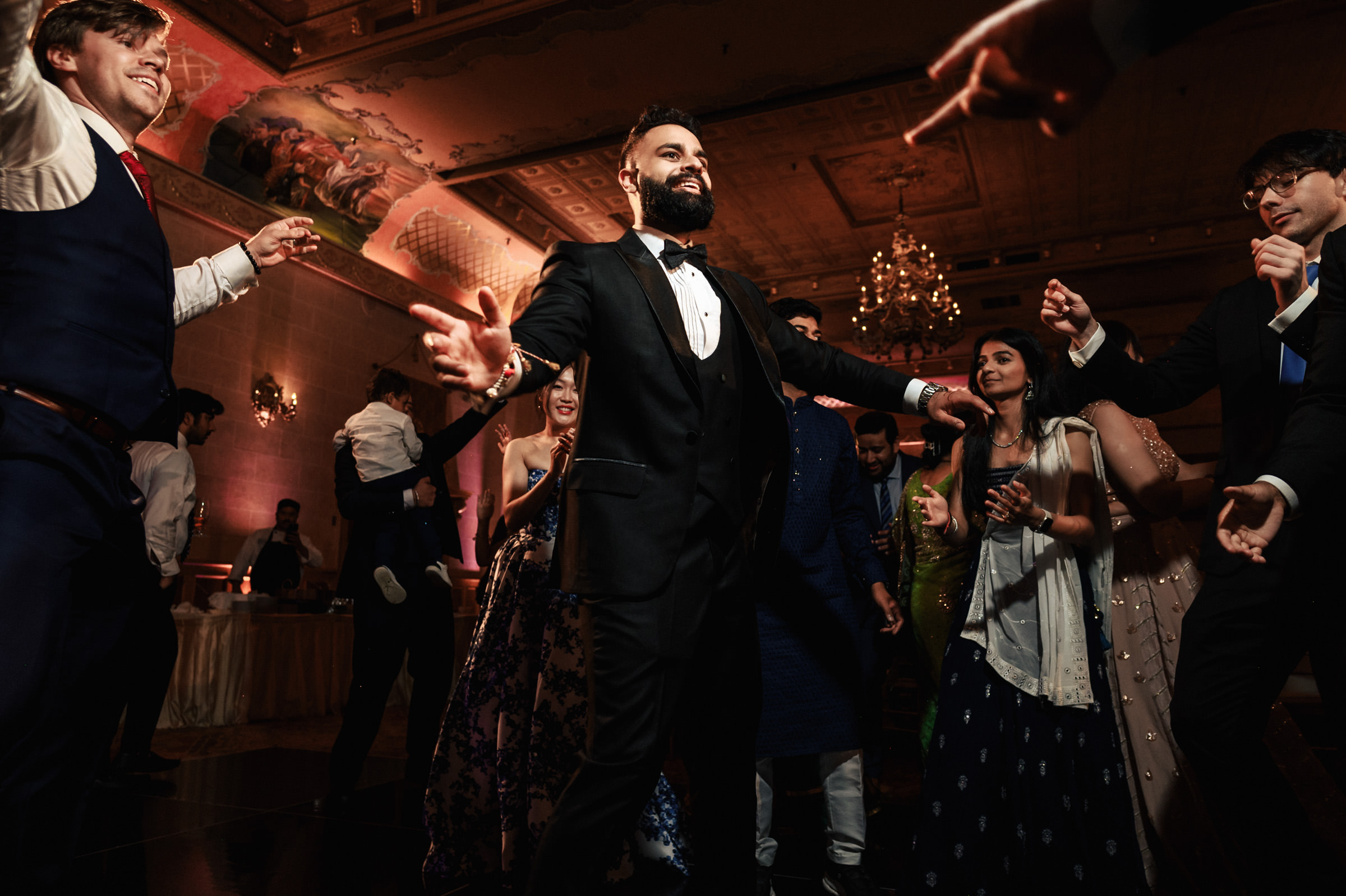 A group of people are dancing and celebrating indoors at an event. The central figure, dressed in a black suit and bow tie, has his arms outstretched. Other participants in formal and semi-formal attire are laughing and dancing around him. The room is dimly lit with chandeliers.