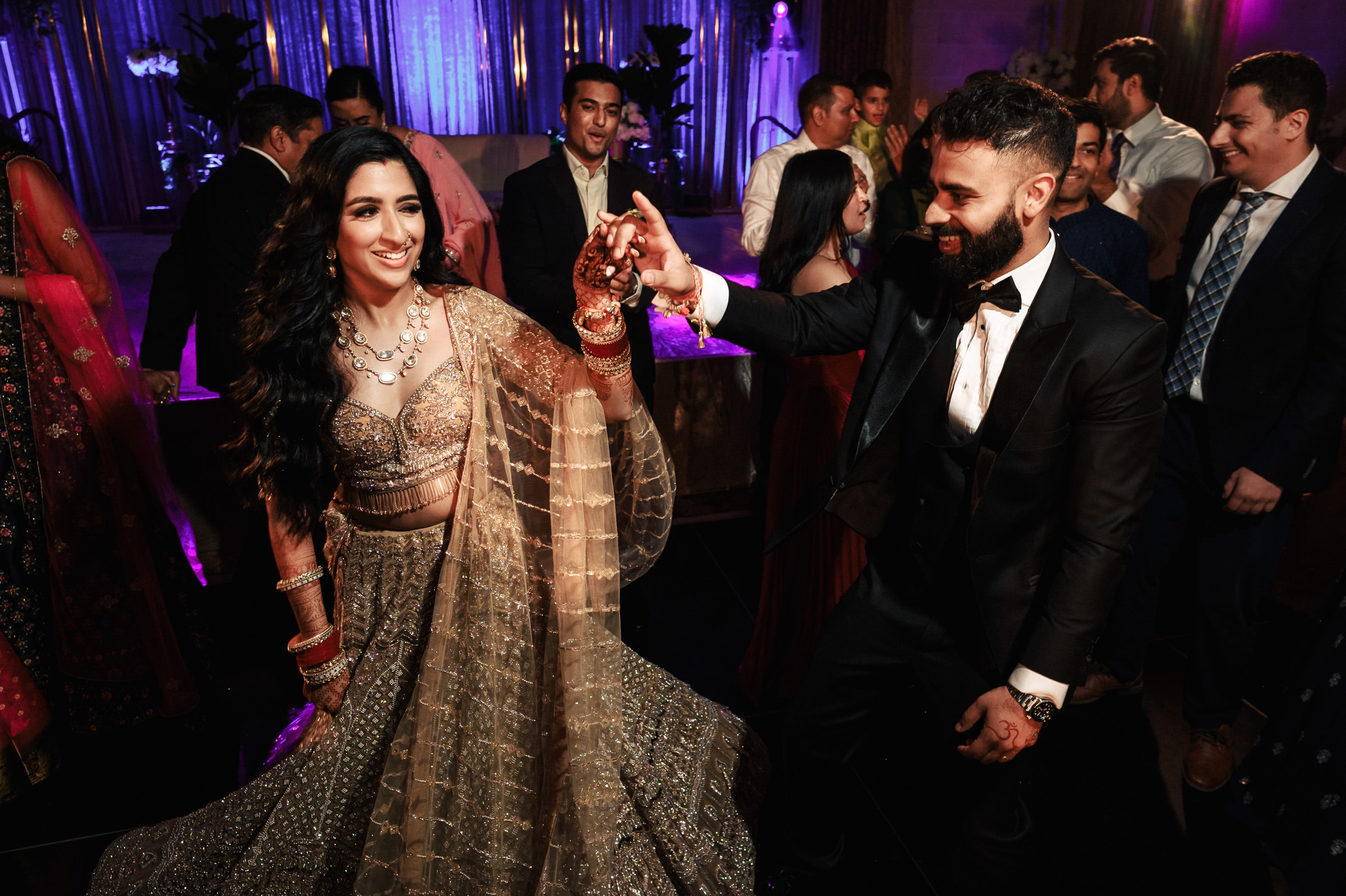 A couple, dressed in formal attire, dances joyfully at a well-decorated evening event. The woman is wearing a glittering traditional dress with intricate jewelry, while the man is in a black suit and white shirt. People in elegant clothing surround them on the dance floor.