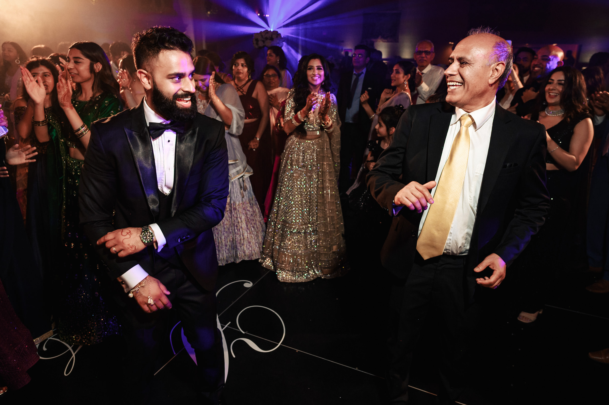 Two men in formal attire, one in a black suit and bow tie and the other in a dark suit with a yellow tie, dance joyfully at a lively social event. Guests in elegant dresses and suits surround them, clapping and smiling, against a backdrop with colorful lights.