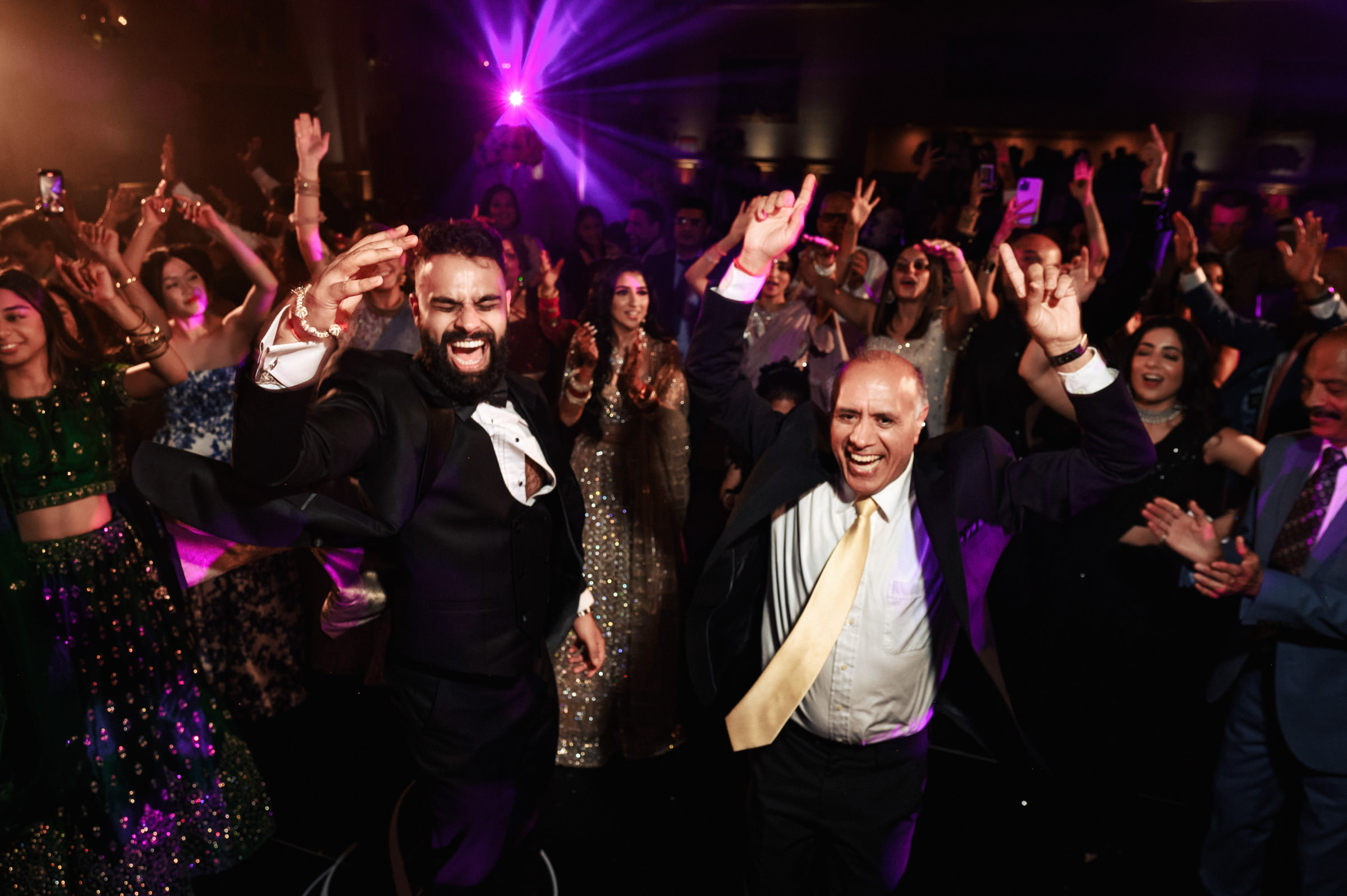 Two men in formal attire, one in a black suit and the other in a black suit with a yellow tie, dance joyfully with their hands in the air at a lively event. They are surrounded by a festive crowd, and colorful lights illuminate the scene, creating a celebratory atmosphere.