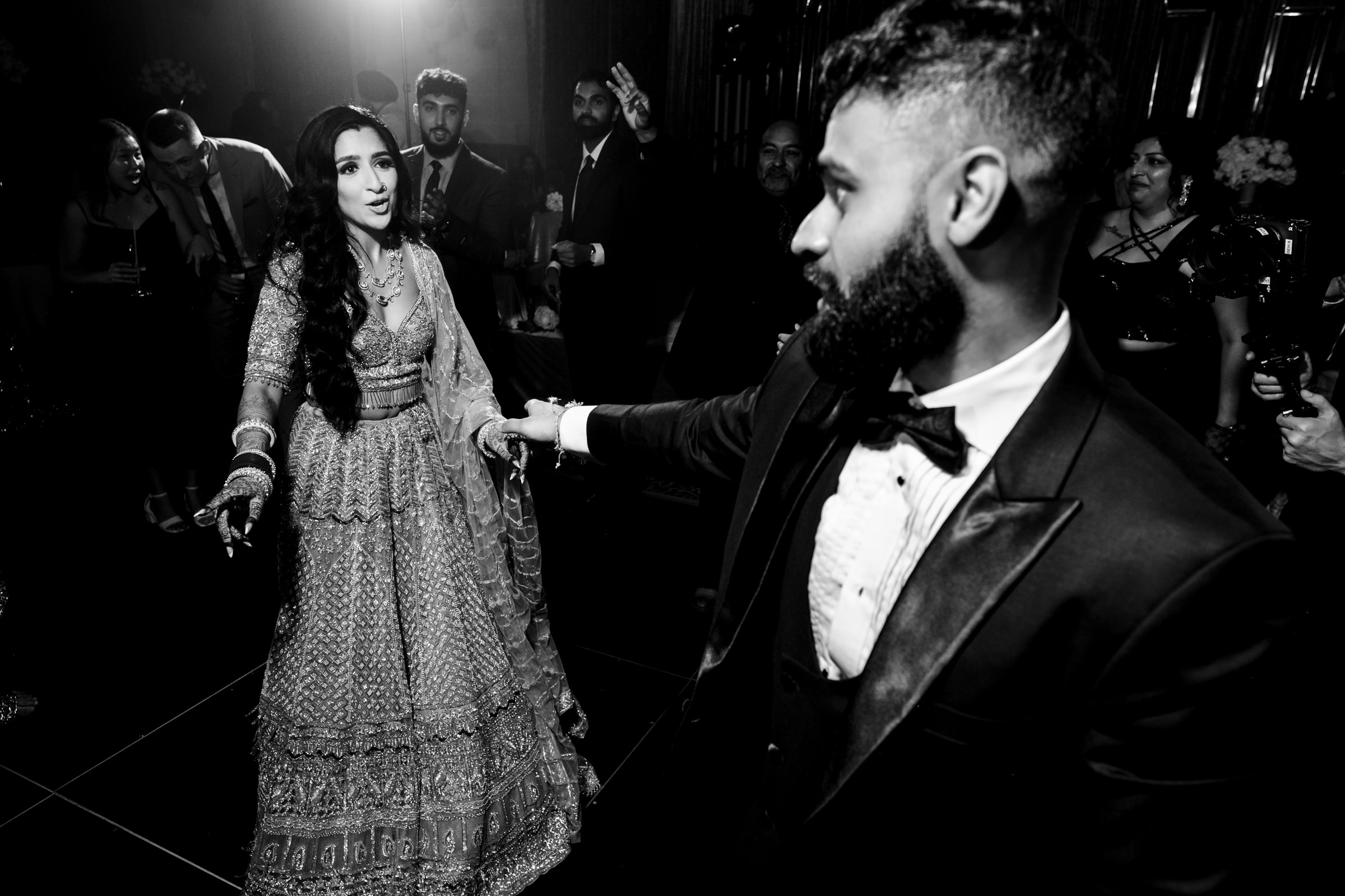 Black and white image of a joyous couple dancing at a celebration. The woman is in a detailed traditional dress, and the man, sporting a beard, is in a formal suit. Guests in the background watch and join in the festivities.