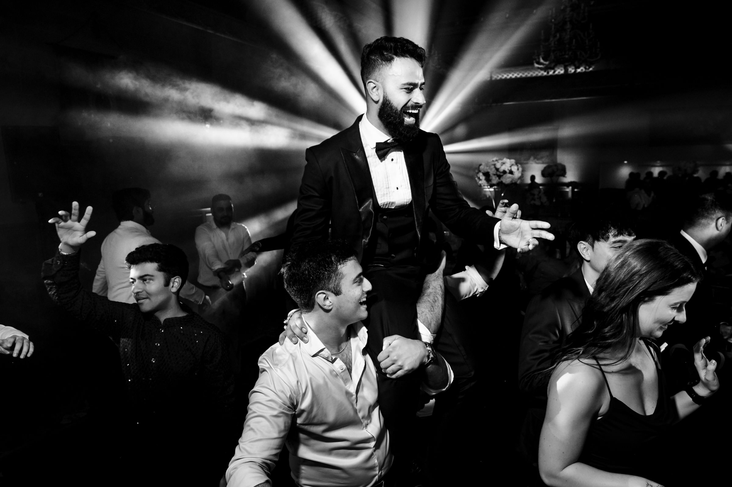 A well-dressed man is being lifted on the shoulders of another man at a lively event. Several people are dancing and cheering around them, illuminated by dramatic stage lighting and beams of light in the background, creating a festive atmosphere.