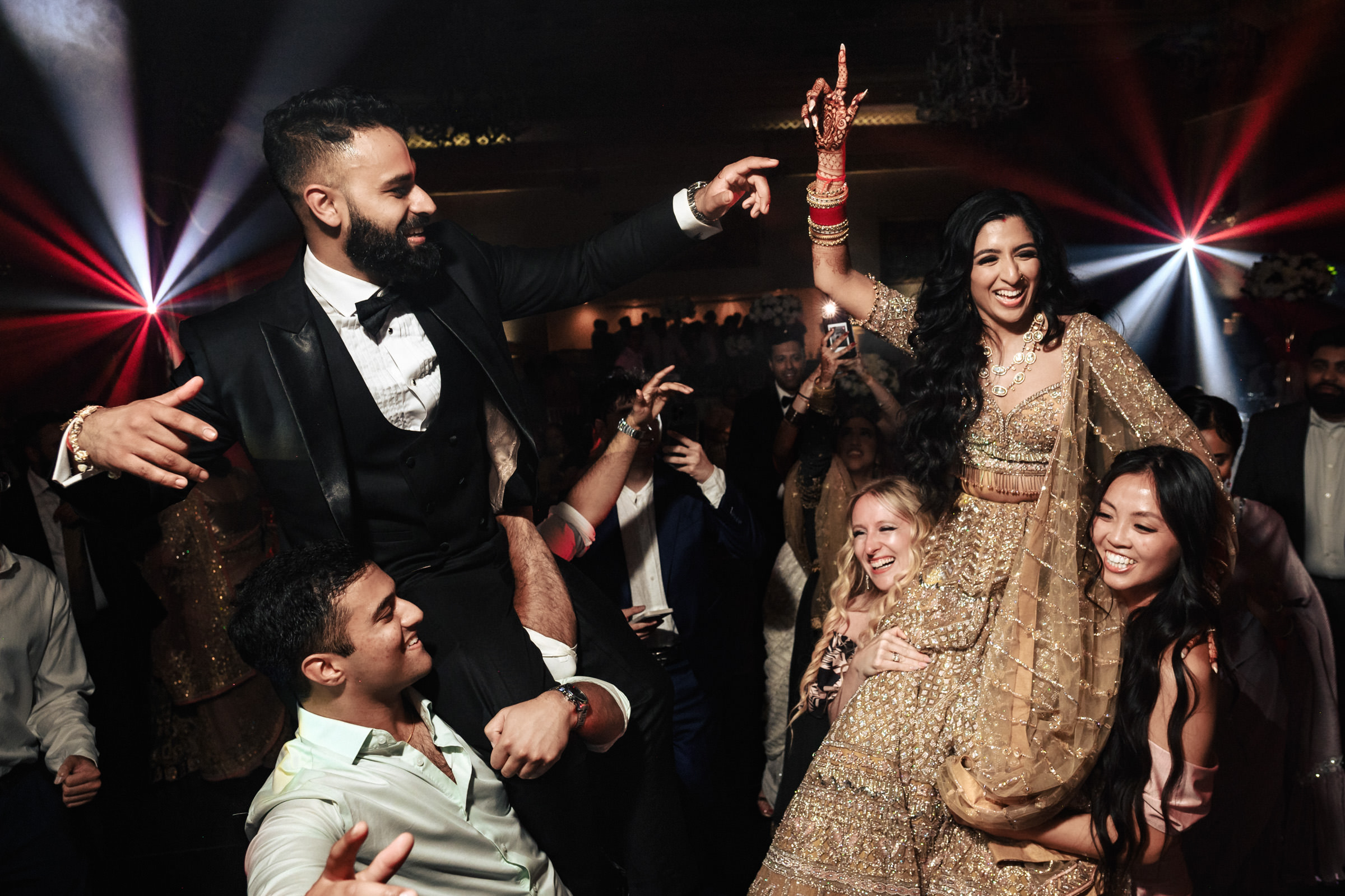 A lively wedding celebration with a bride in a gold dress and a groom in a black suit lifted on the shoulders of guests. The bride raises her hand joyfully while surrounded by smiling friends. Colorful lights and joyful expressions enhance the festive atmosphere.