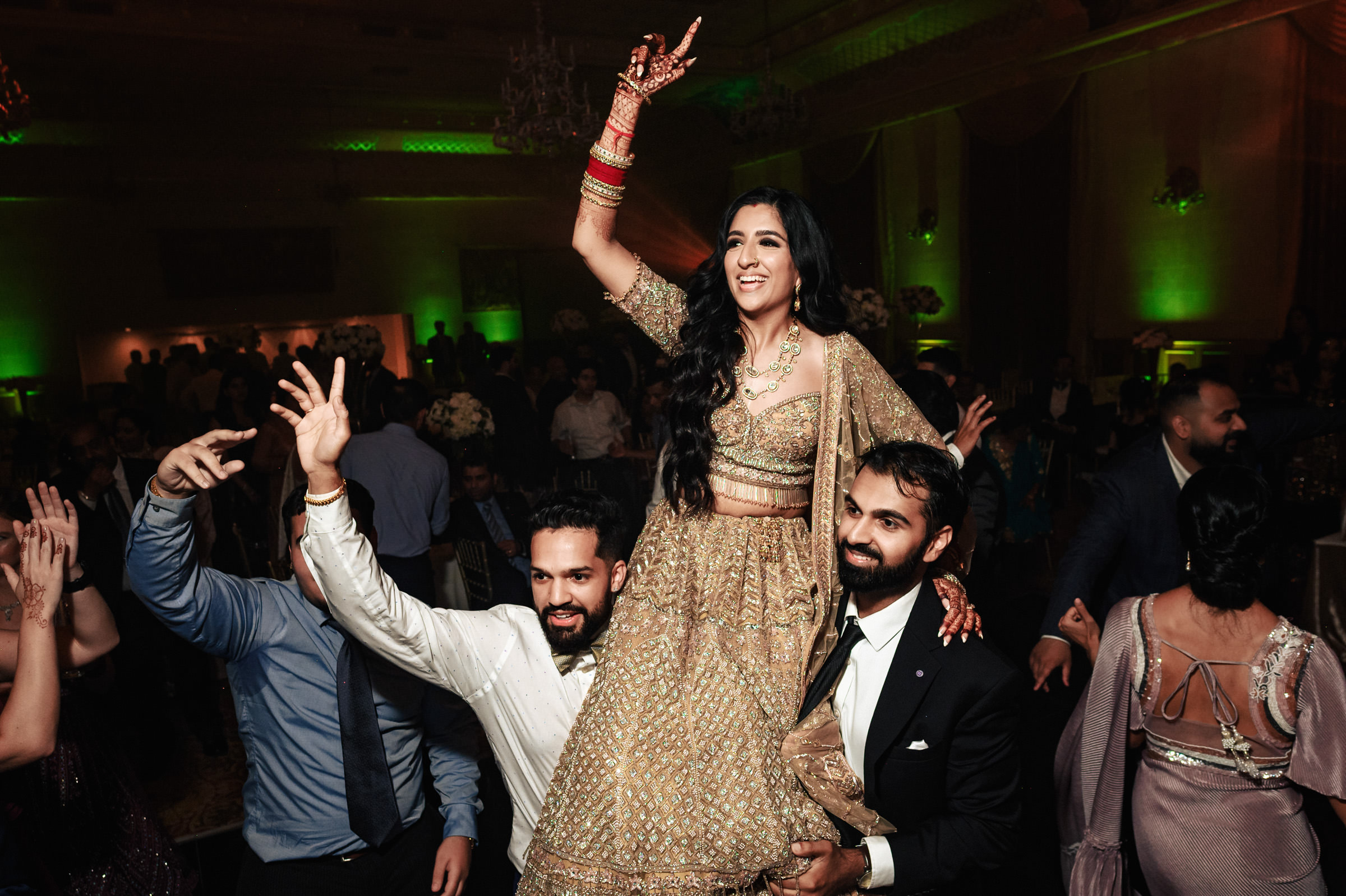 A woman in an ornate traditional outfit is lifted by two men, one on each side, as she dances and smiles joyfully at a festive event with dim lighting and green accent lights. The crowd around them is cheering and dancing.
