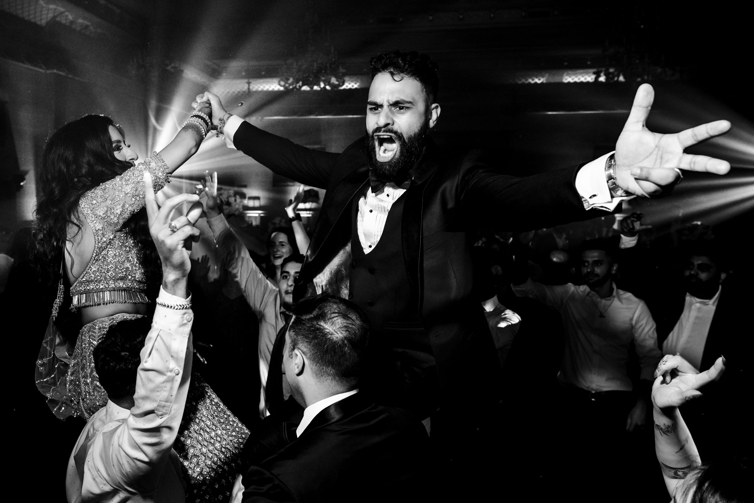 Black and white photo of a lively celebration. A man in a tuxedo with arms outstretched and an ecstatic expression is at the center. Around him, people are cheering, dancing, and supporting a woman lifted in the air, also in festive attire. Spotlights and energy fill the scene.