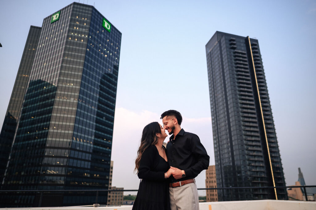 A couple stands close together on a rooftop, smiling and holding hands. Tall, modern skyscrapers with reflective windows dominate the background. The sky is clear with a hint of dusk light, adding a warm glow to the scene.