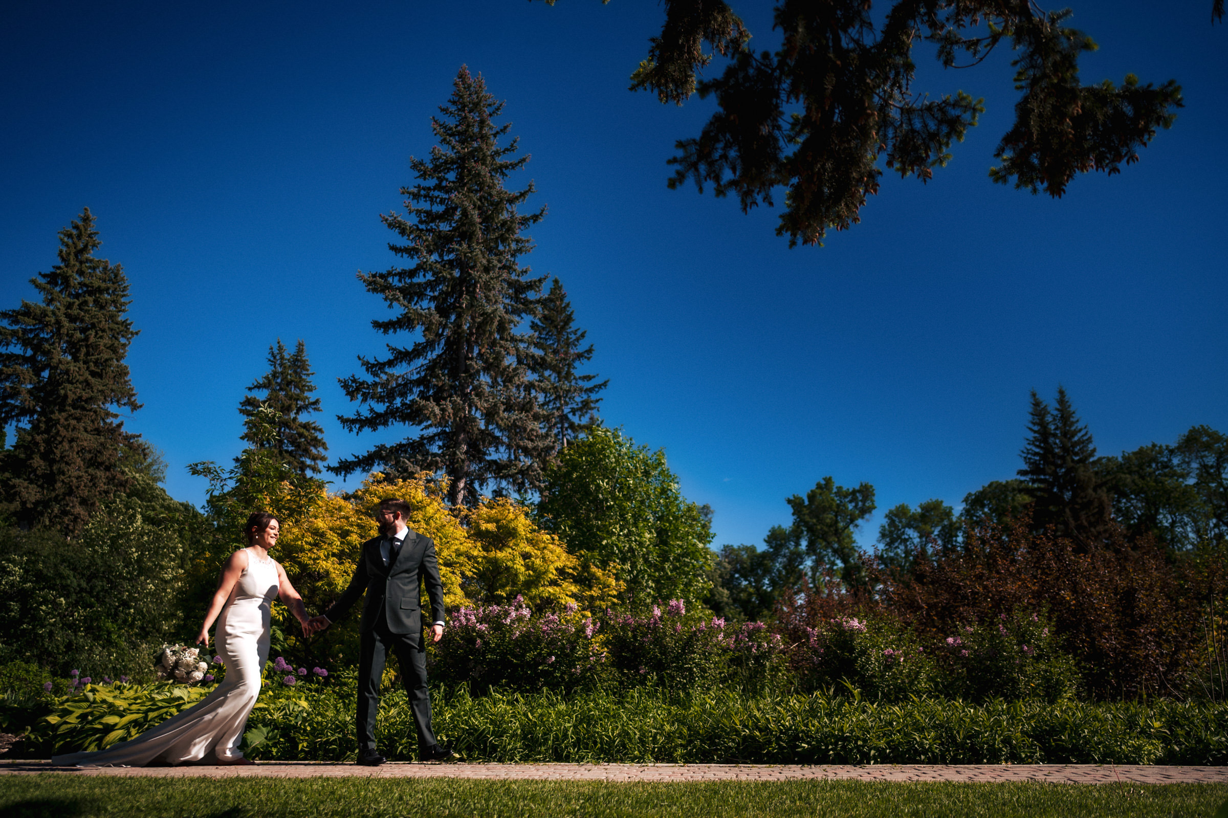Bride and groom stroll hand in hand at a Winnipeg wedding venue under a clear blue sky.