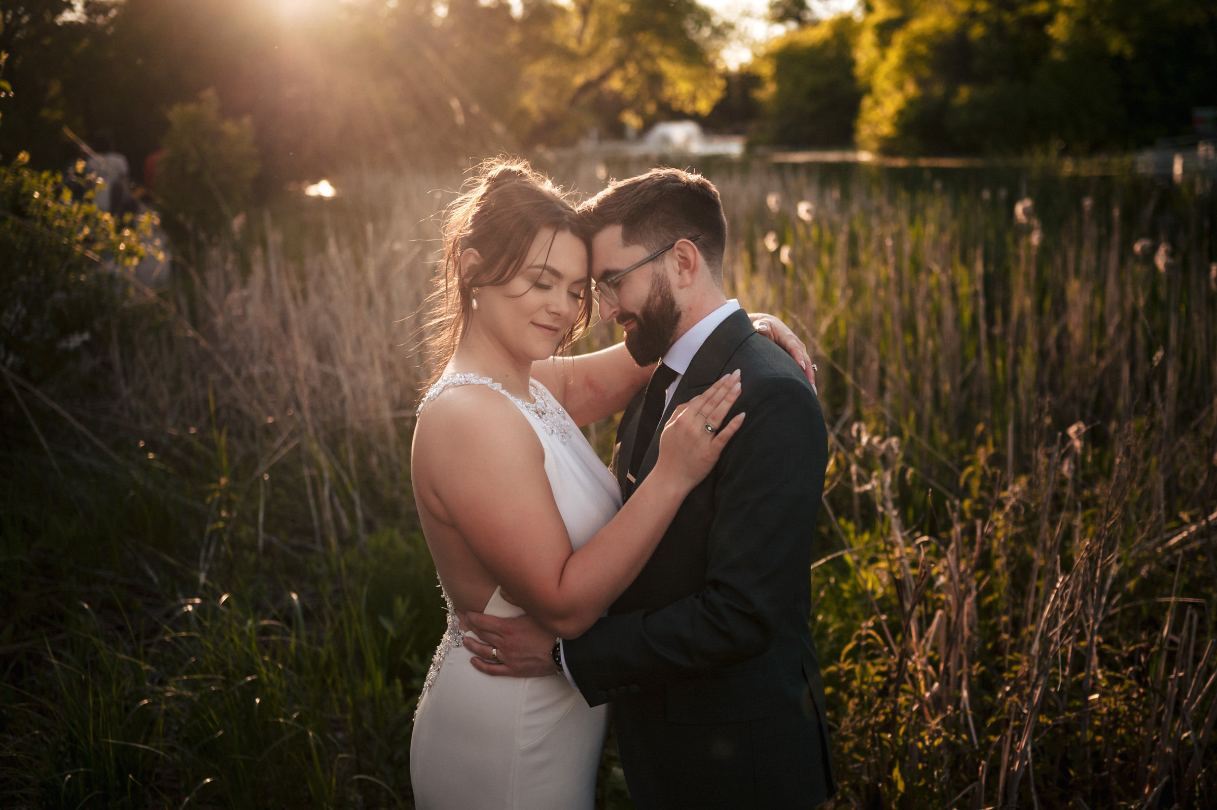 Bride and groom embracing in a sunlit Winnipeg wedding venue with tall grass and trees.