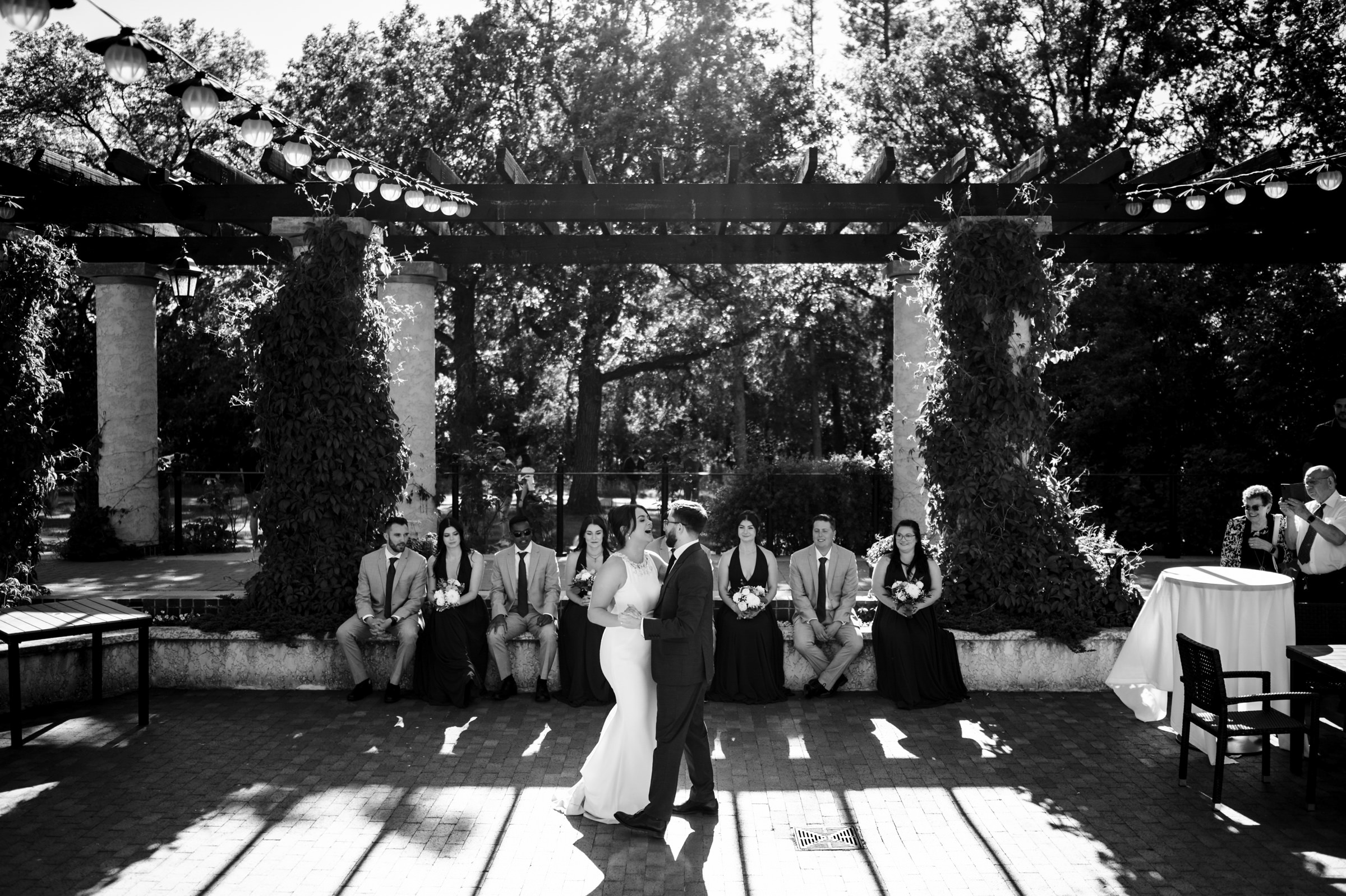 Bride and groom dancing outdoors at a Winnipeg wedding venue, bridal party under a pergola.