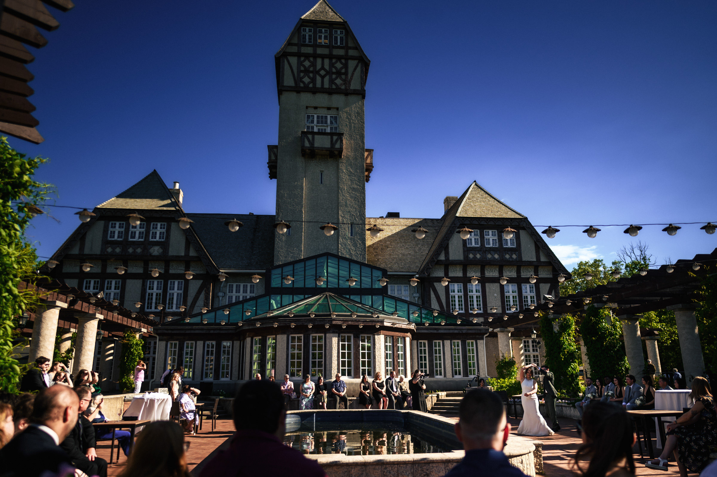 Outdoor wedding by a historic Winnipeg venue with a towering spire and clear blue sky.