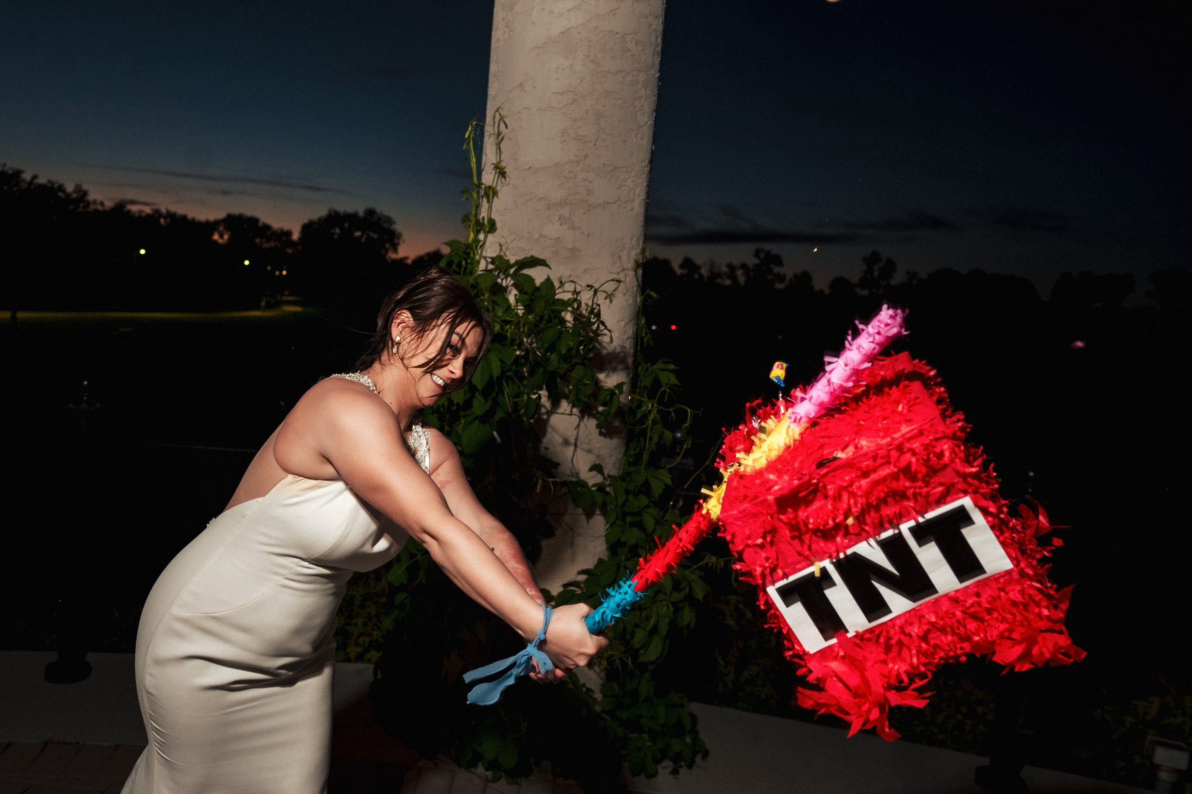 Person in a white dress hitting a red "TNT" piñata outdoors at a Winnipeg wedding venue.