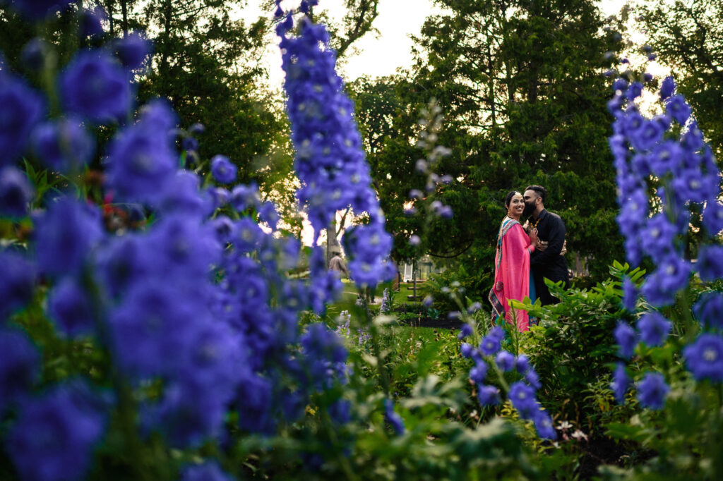 A couple standing closely together and smiling in a lush garden. The foreground features large, vibrant purple flowers in sharp focus, while the couple and greenery in the background are slightly blurred. Tall trees are visible in the distance.