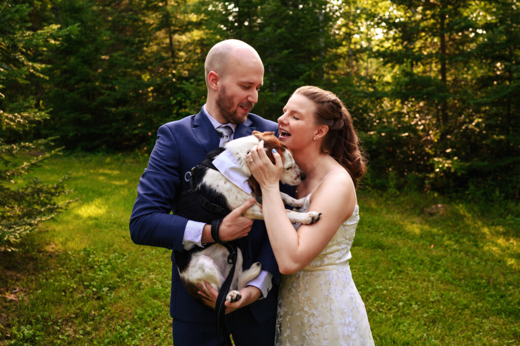 A couple stands outdoors in a grassy, wooded area. The man, dressed in a suit, holds a dog in his arms. The woman, in a white dress, smiles and gently touches the dog's face. Sunlight filters through the trees, creating a warm atmosphere.