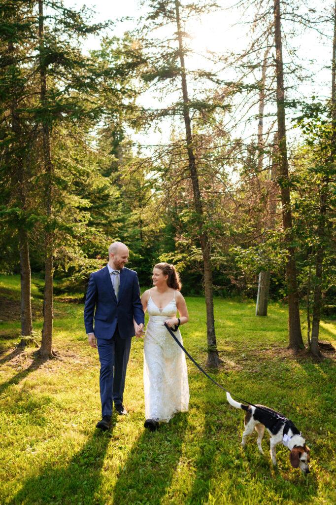 A couple walks hand in hand through a sunlit forest, both dressed in formal attire. The woman wears a wedding dress, and the man is in a suit. They are accompanied by a beagle dog on a leash. Sunlight filters through the trees, casting gentle shadows.