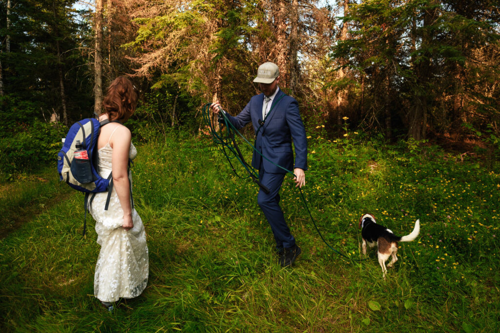 A person in a wedding dress with a backpack stands on grass, watching another person in a suit with a cap as they walk a dog on a leash. They are surrounded by lush green trees and plants, with sunlight filtering through the branches.