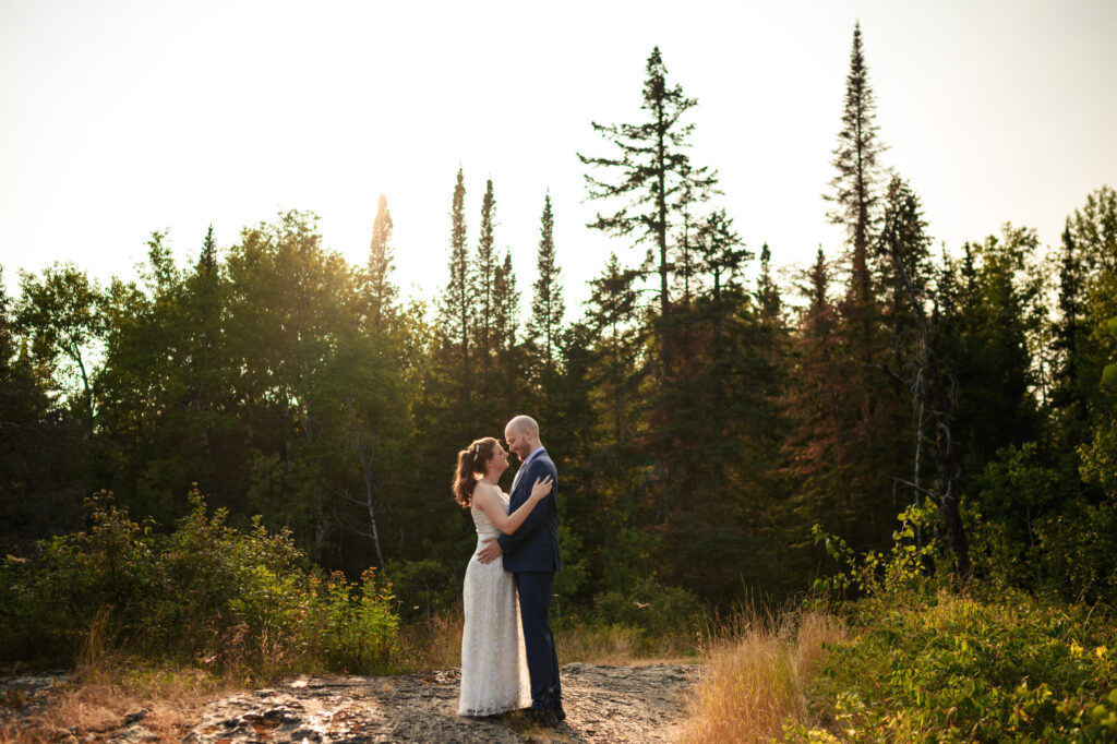 A couple embracing on a sunlit, grassy landscape with tall trees in the background. The bride is in a white dress, and the groom is in a dark suit. The sunset creates a warm, tranquil atmosphere.