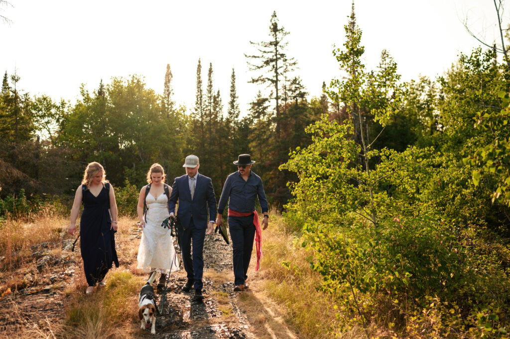 A bride and groom walk along a forest path with two companions and a dog. Sunlight filters through the trees, casting a warm glow over the scene. The group appears relaxed and joyful, enjoying the natural surroundings.