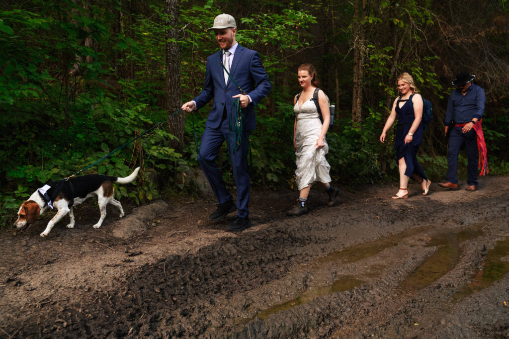 A group of four people walks along a muddy trail in a forest. The person in front, wearing a suit and cap, leads a dog on a leash. The others follow, dressed in formal attire. Lush green foliage surrounds them.