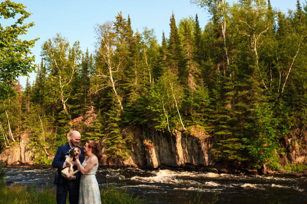 A couple stands by a flowing river surrounded by dense green forest. They are smiling and holding a dog. The sky is clear and the sunlight filters through the trees, creating a serene and joyful atmosphere.