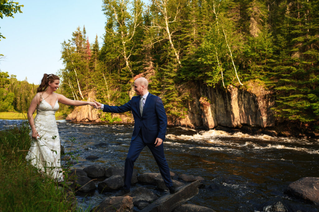 A couple stands by a forest river; the woman in a white dress holds the hand of a man in a navy suit. She stands on rocks near the water, and he assists her. Lush green trees are in the background under a clear sky.