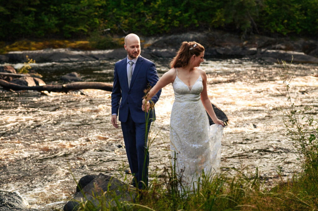 A couple in formal attire walks along a riverbank. The woman, in a white dress, holds her skirt as they move through the grass. The man, in a blue suit, follows closely. The river flows rapidly in the background, surrounded by lush greenery.