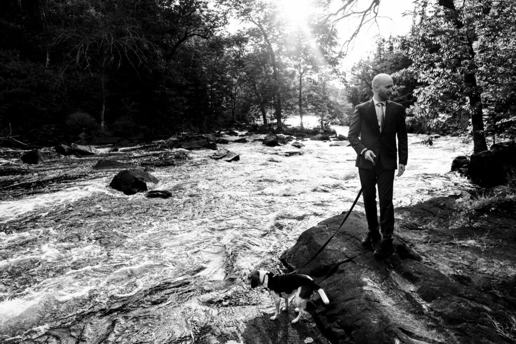 A man in a suit stands on a rock by a flowing river, holding a leash for a small dog. Sunlight filters through trees, casting rays across the water. The scene is in black and white, highlighting the contrast between the dark woods and bright river.