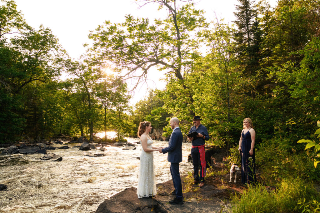 A couple stands on a rock by a river exchanging vows in an outdoor wedding. A woman and an officiant stand nearby, with a small dog beside them. The scene is surrounded by lush green trees and bathed in warm sunlight.