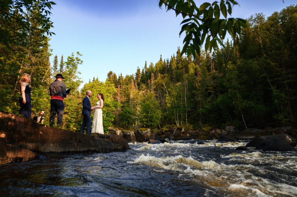 A wedding ceremony beside a flowing river in a forested area. The bride and groom stand on a rocky edge while an officiant and a guest watch. Sunlight filters through the trees, illuminating the scene with a warm glow.