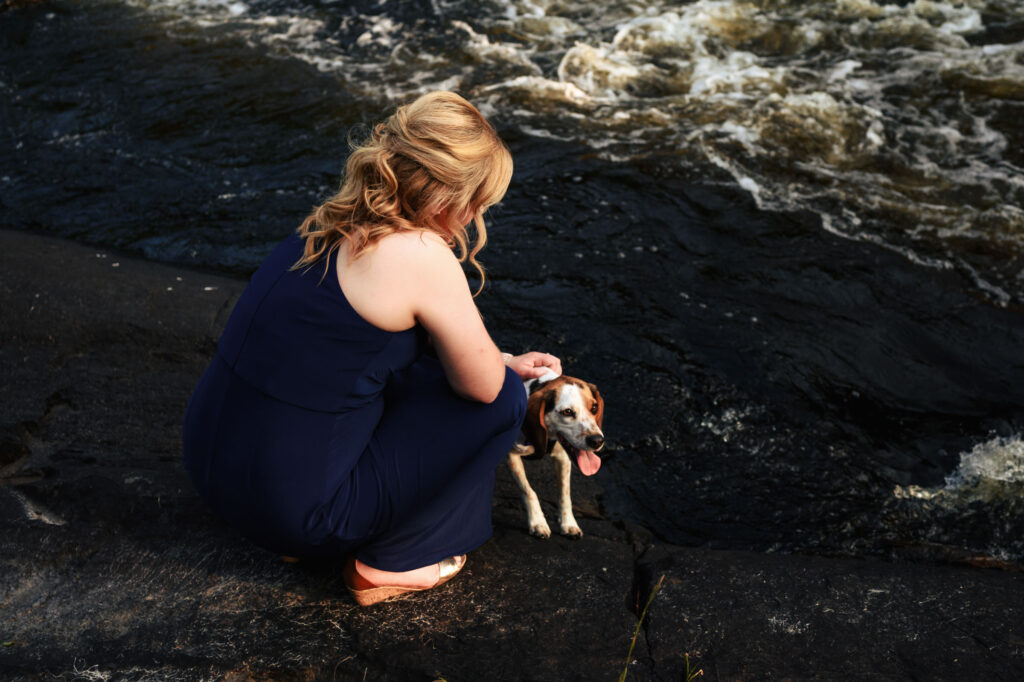 A woman with blond hair in a blue dress crouches by the edge of a flowing river, gently petting a small dog with its tongue out. They are both on a stone or concrete surface by the water.