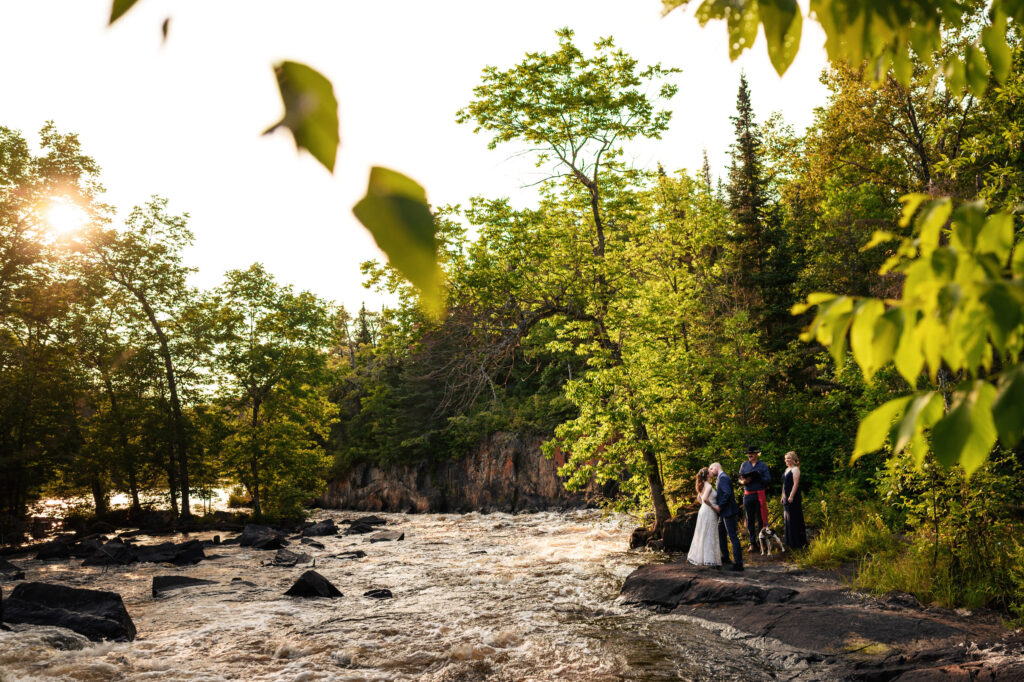 A couple is getting married by a flowing river surrounded by lush green trees. The sun is setting, casting a warm glow over the scene. A few guests are present, standing on the rocks by the water.